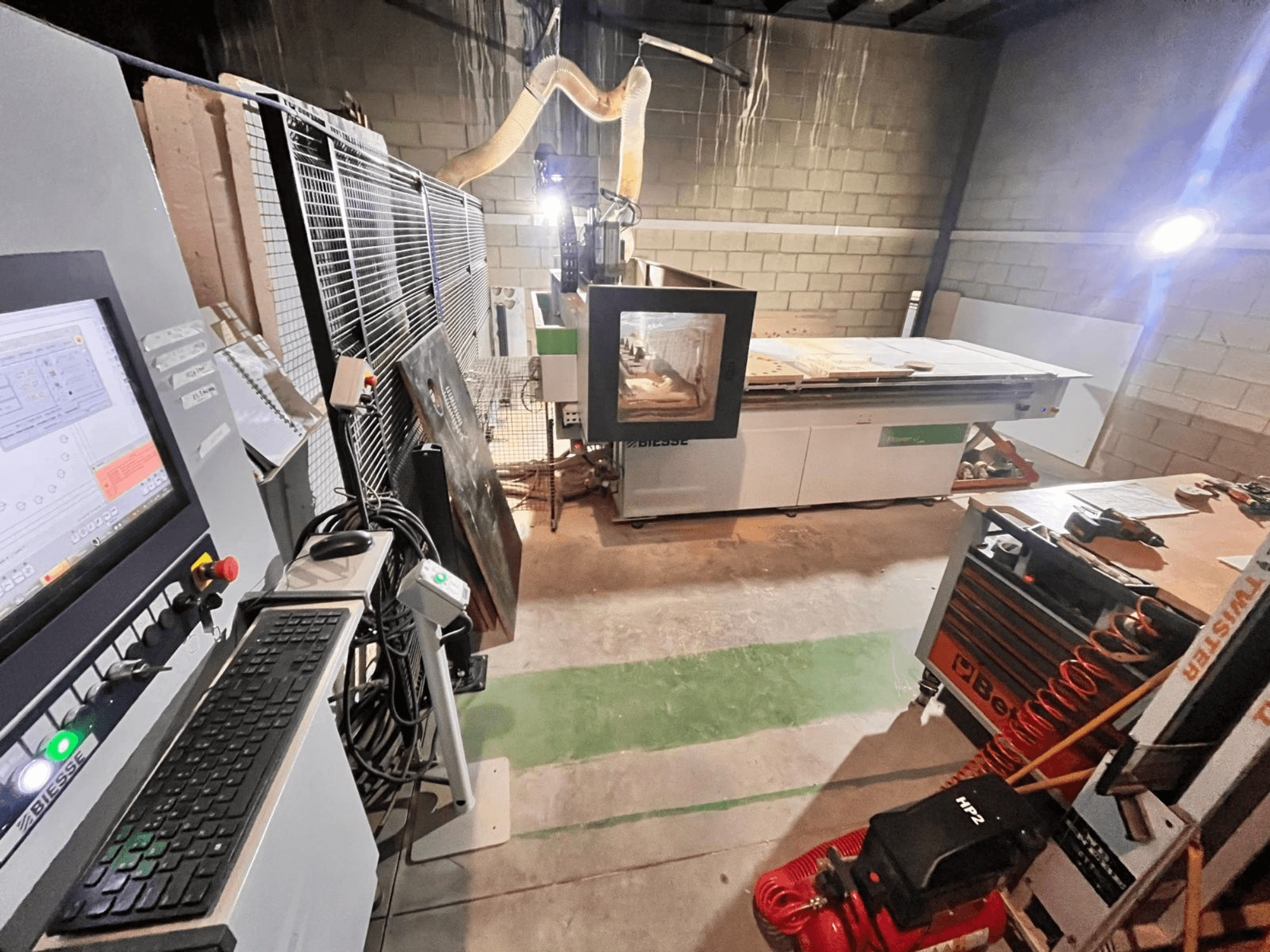 Atölyedeki Biesse CNC makinesi, kontrol paneli, toz emme, aletler ve tabla üzerindeki çeşitli malzemeleri gösteren yandan görünüm.