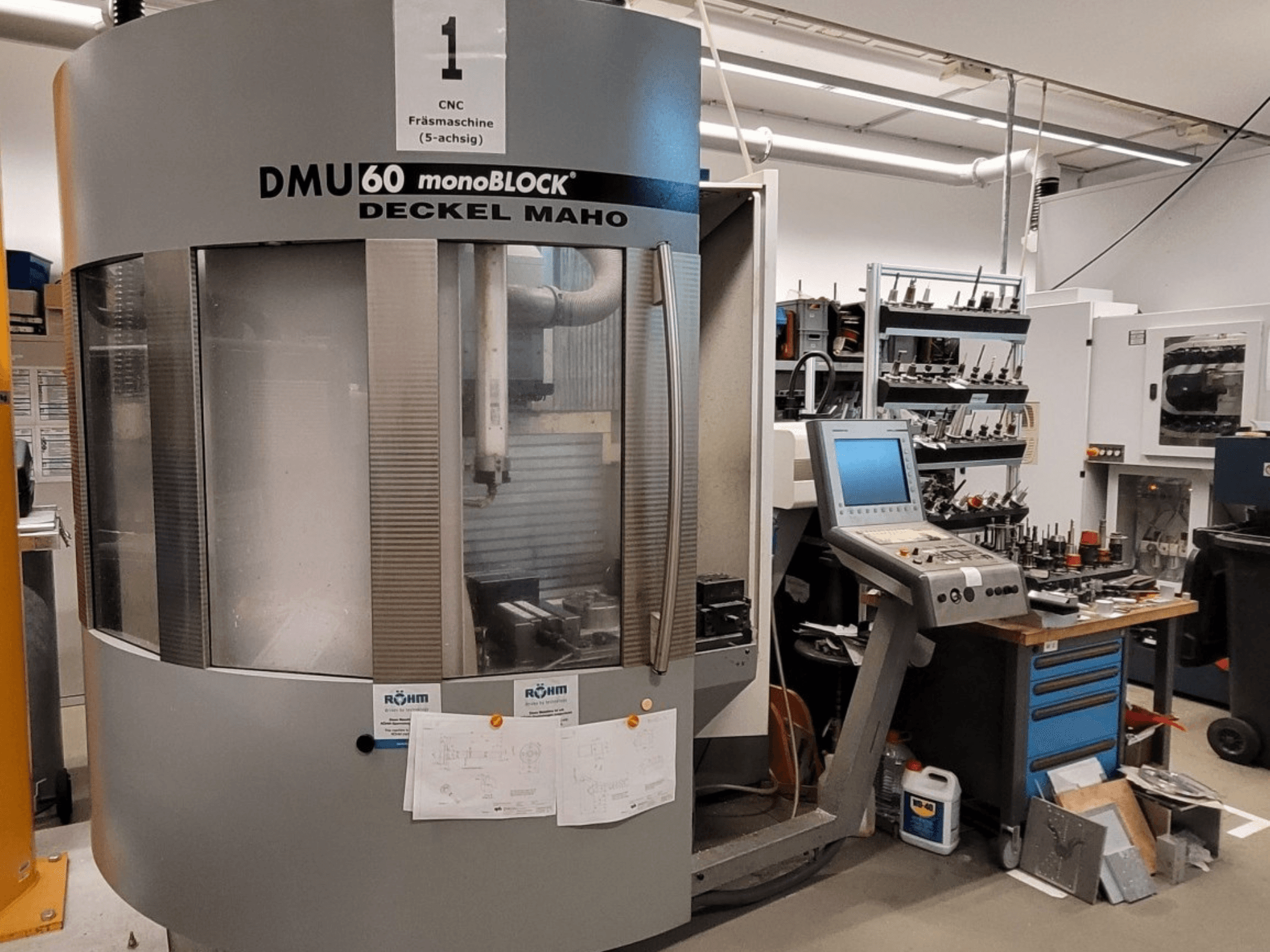 CNC freze makinesi DECKEL MAHO DMU 60 monoBLOCK önden görünümde, kontrol paneli ve yanındaki takım deposu sergileniyor.