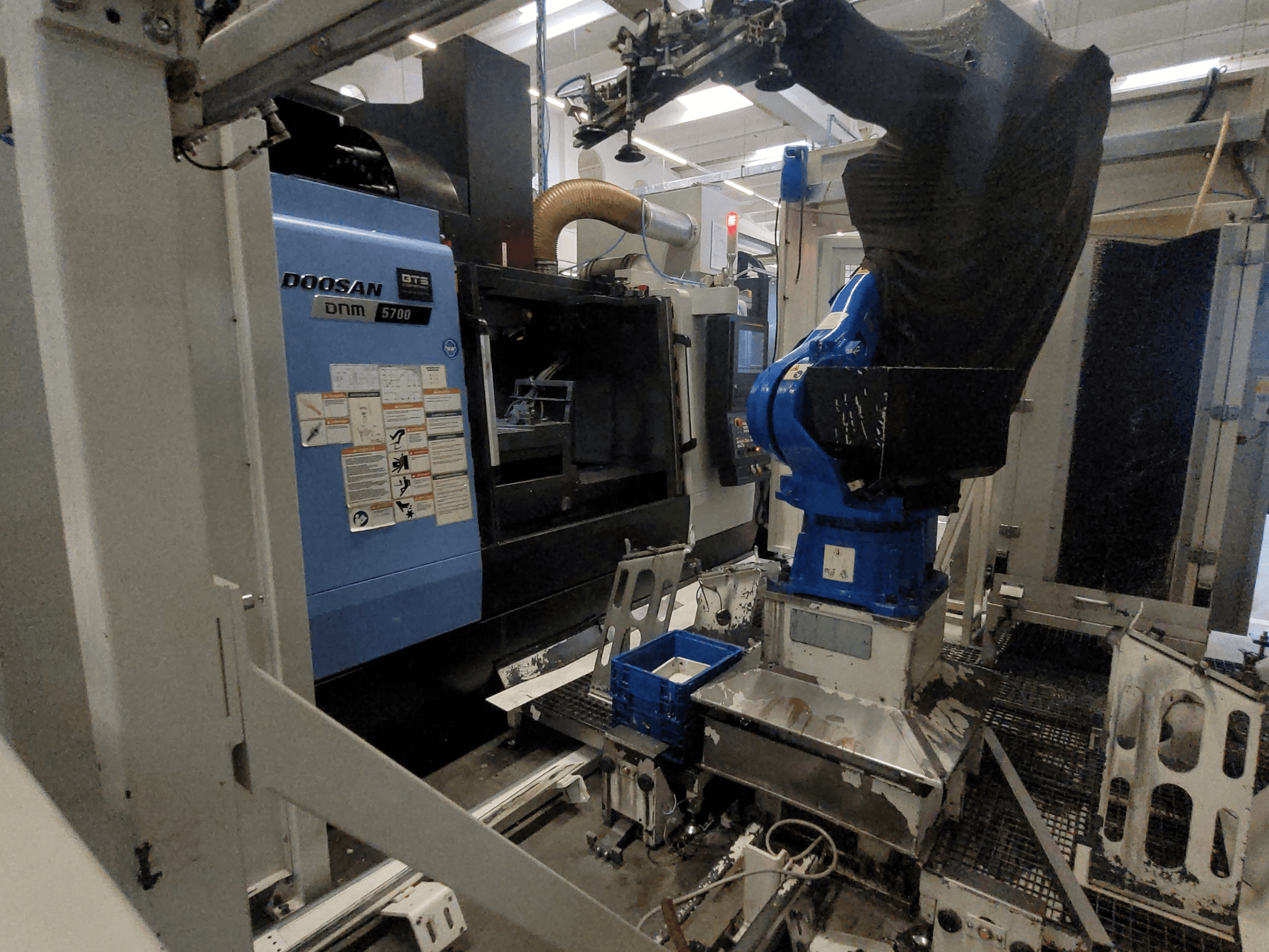 Fabrika ortamında çekilmiş DOOSAN DNM 5700 dikey işleme merkezinin ön görünümü ve robot kol entegrasyonunu gösteren fotoğraf.