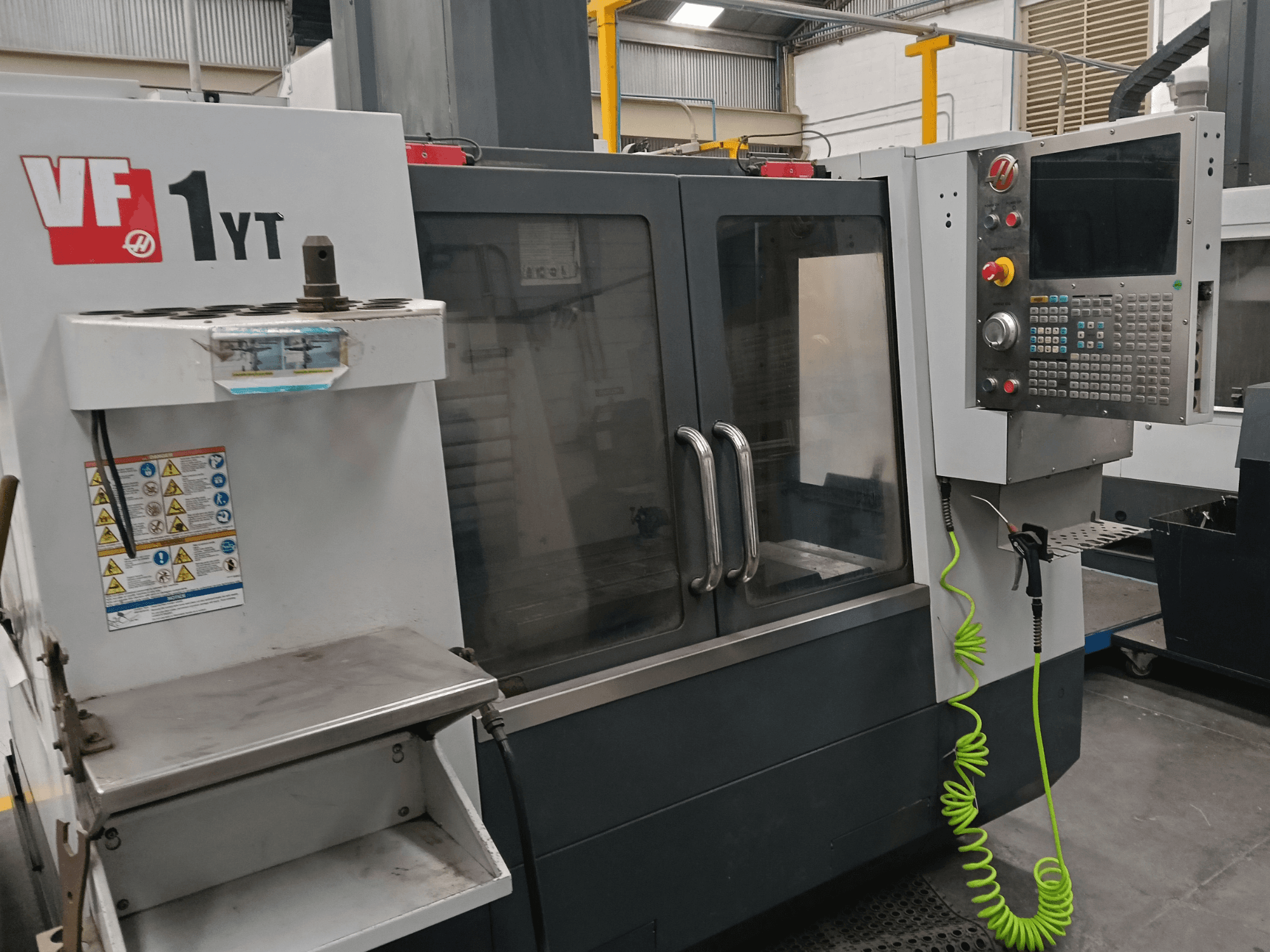 HAAS VF-1YT-2 Dikey İşleme Merkezi, önden görünüm; kontrol paneli, takım tutucu, güvenlik etiketleri ve işleme alanı gösterilmektedir.