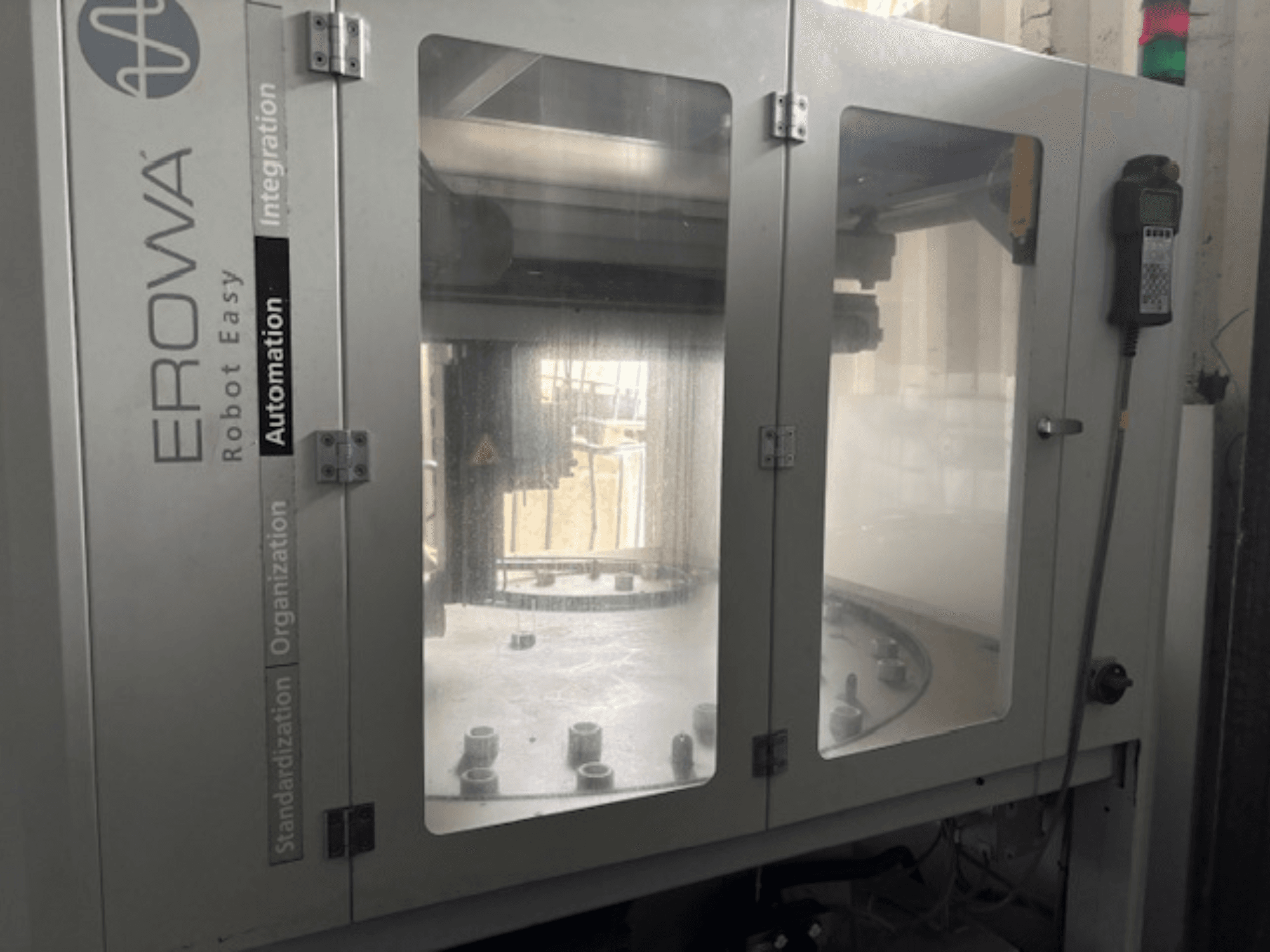 EROWA Robot Easy Automation'ın önden görünümü; şeffaf kapılar, görünür otomasyon bileşenleri ve kontrol paneline sahiptir.