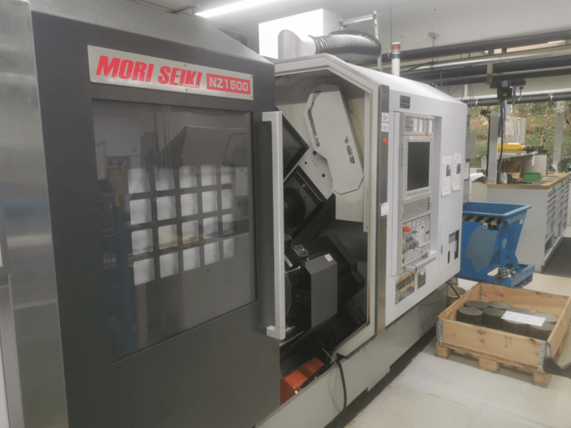 MORI SEIKI NZ1500 CNC makinesi hafif bir açıyla, ahşap bir palet üzerinde metal iş parçaları ile kontrol panelini ve çalışma alanını göstermektedir.
