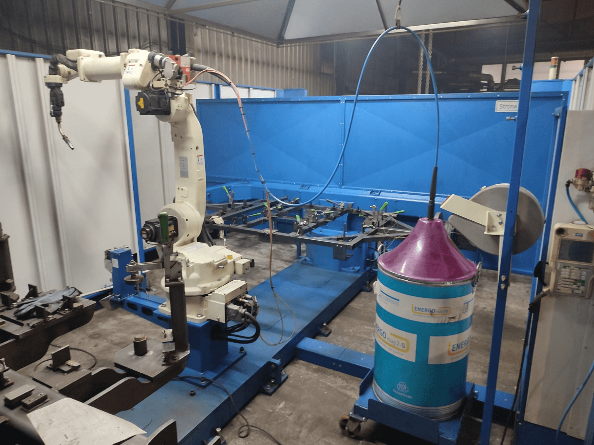 Beyaz ve mavi tabanlı endüstriyel robot kol, mavi bir çalışma yüzeyi ve metalik bileşenlerle bir atölyede kurulmuştur.