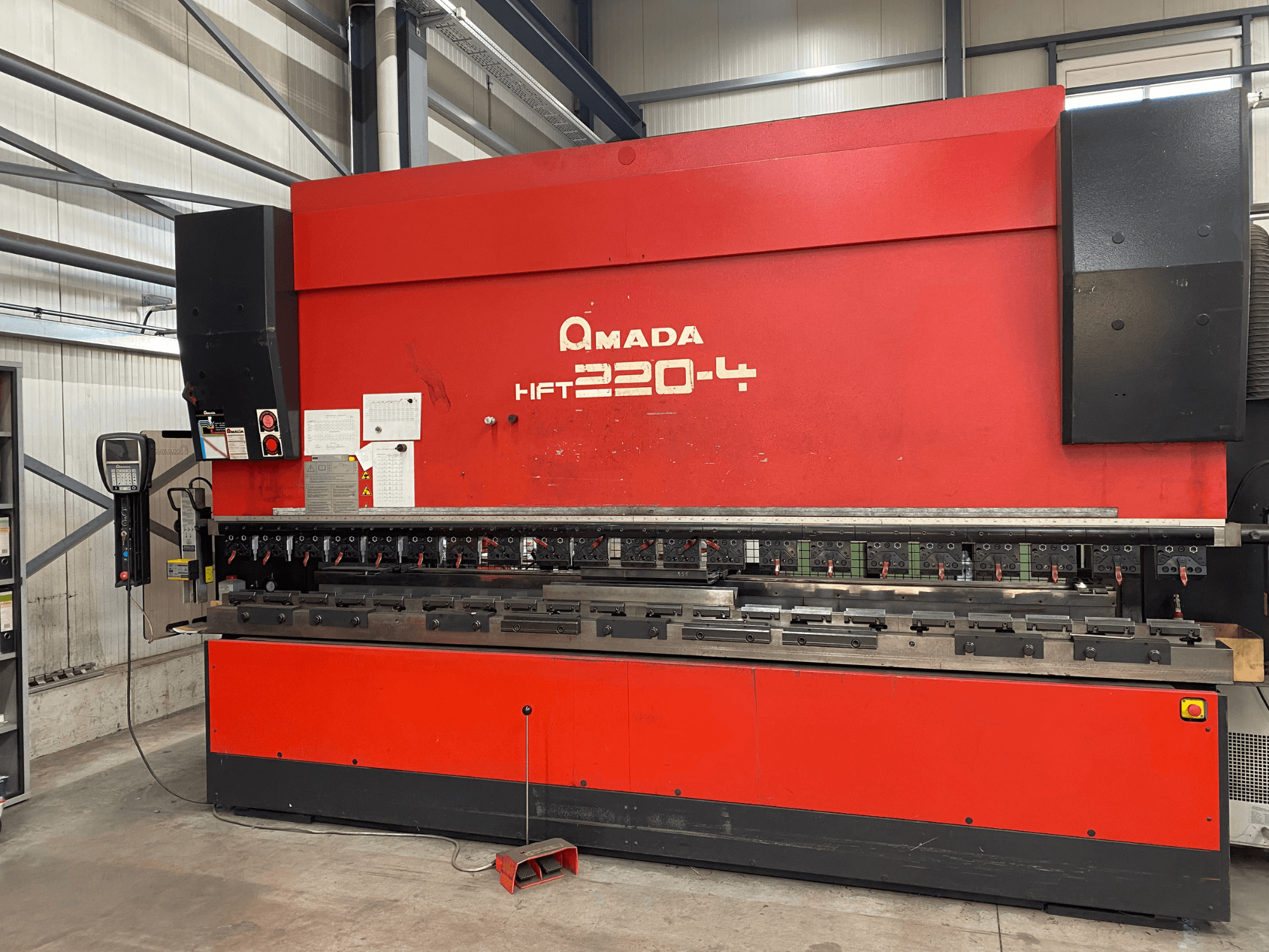 Fabrika ortamında kırmızı AMADA HFT 220-4 abkant pres, kontrol panelini ve çalışma yüzeyindeki bükme aletlerini gösteren önden görünüm.