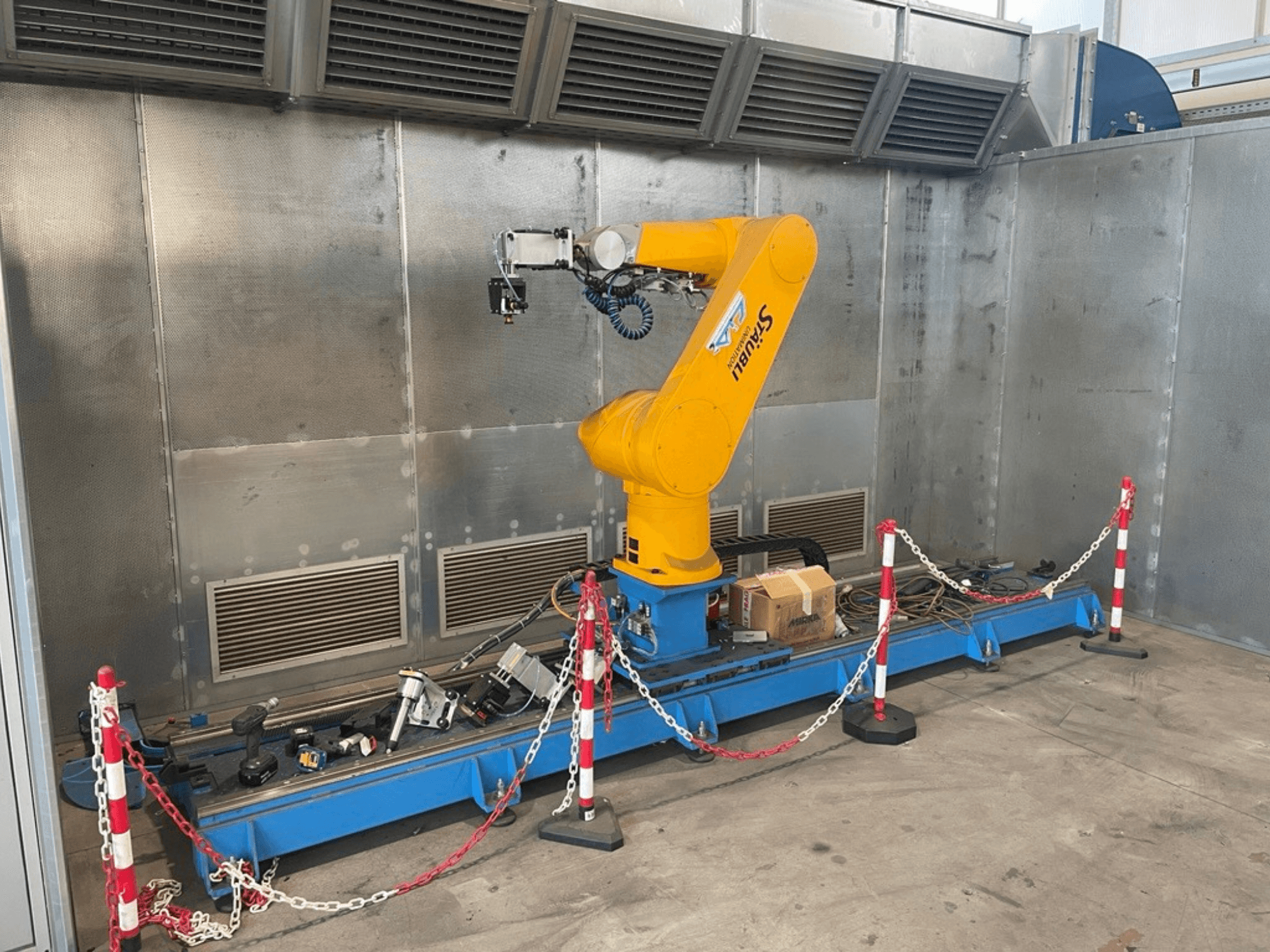 STAUBLI CS8C-RX160 robot kolu bir atölyede, yandan görünüm, koruyucu bariyerlerle çevrili mavi bir taban üzerinde kablolar ve çeşitli aletlerle.