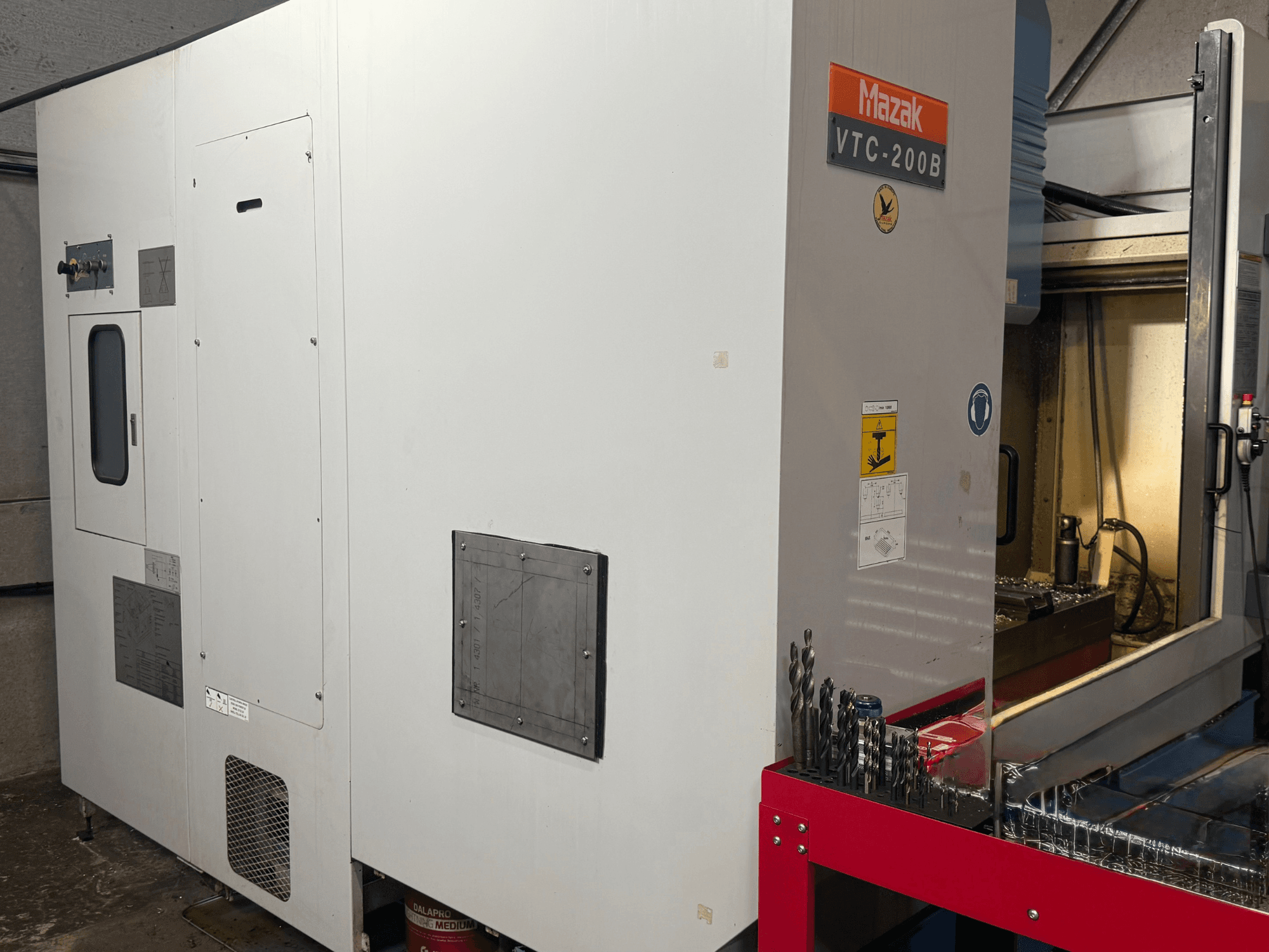 Mazak VTC-200B dikey işleme merkezinin yandan görünümü; etiketli kumanda düğmeleri ve kırmızı saklama rafındaki matkap uçları görülmektedir.