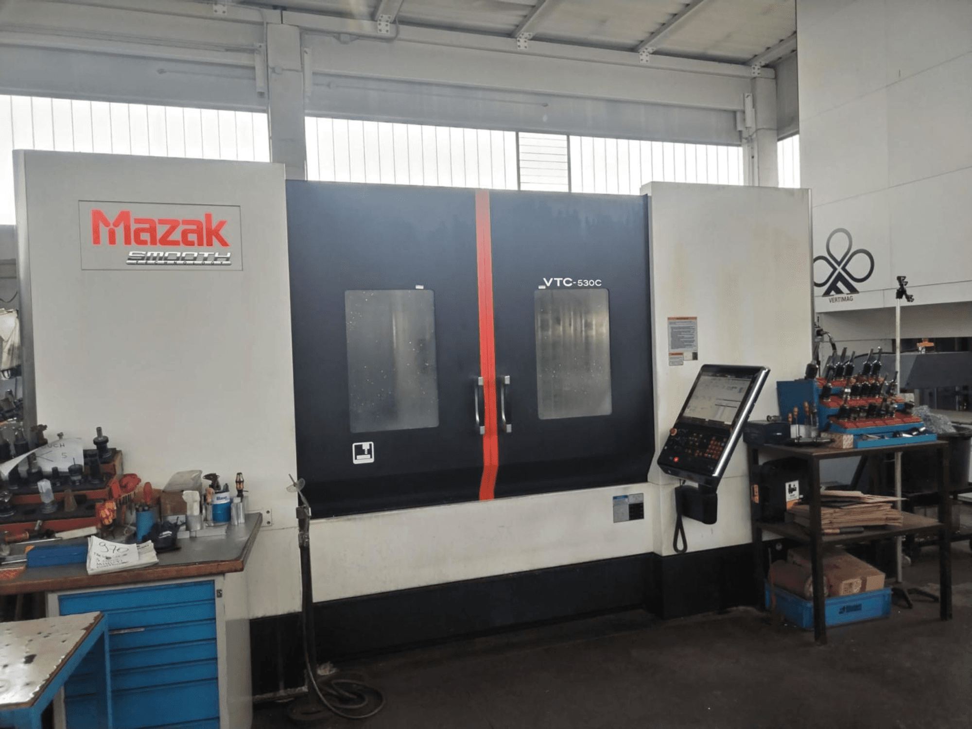 Bir atölyede bulunan, yan masasında kontrol paneli ve aletler bulunan Mazak VTC-530C dikey işleme merkezi.