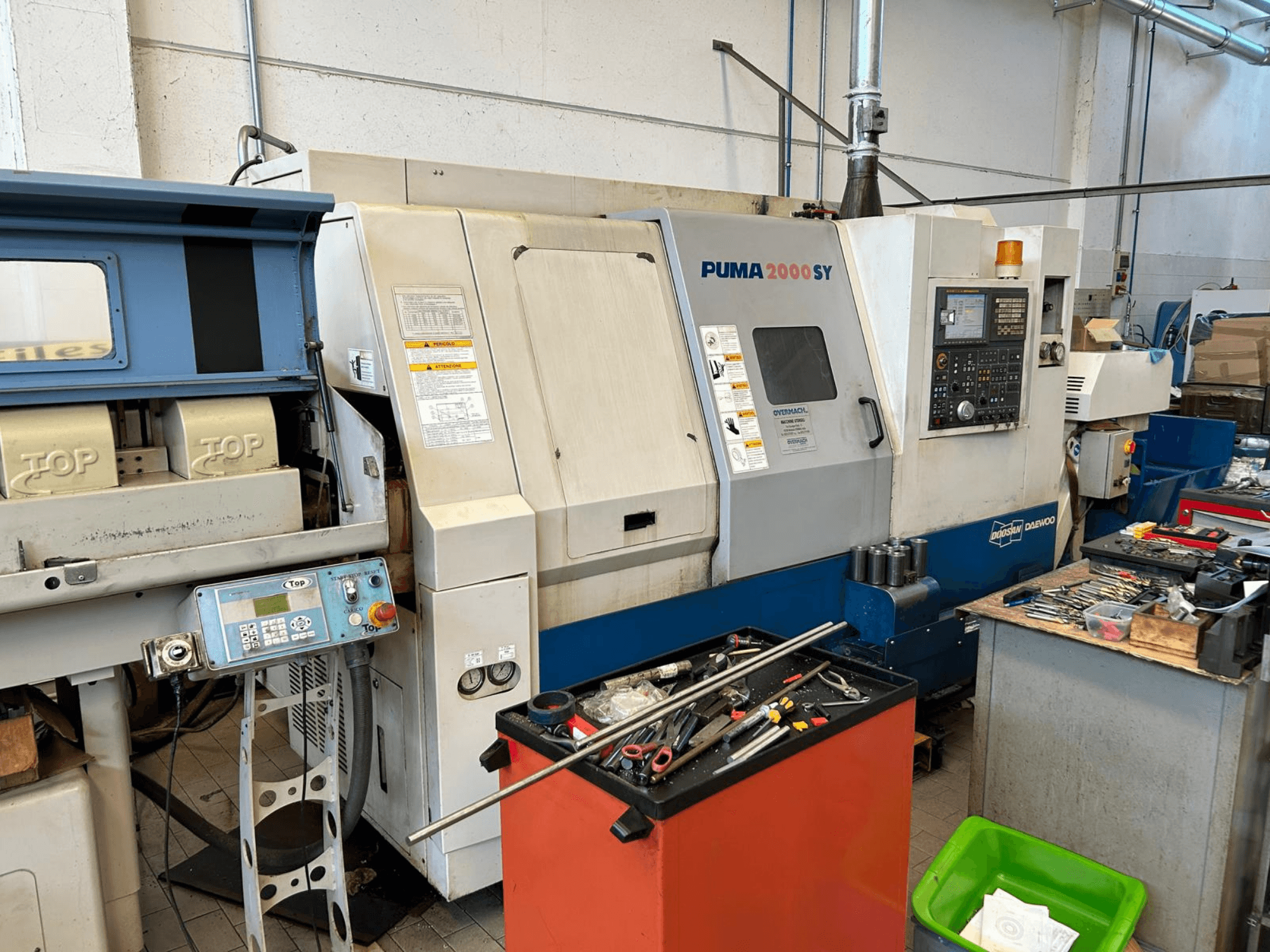 Bir atölyede bulunan DOOSAN PUMA 2000SY yatay torna tezgahı; kontrol paneli ve yanındaki aletlerin görüldüğü yandan görünüm.