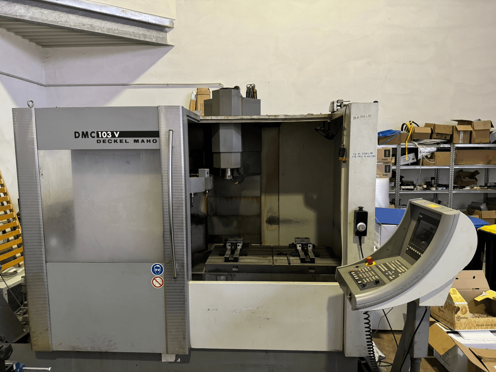 DMC 103 V Deckel Maho freze makinesi, önden görünüm, çalışma panelini, metal parçaları ve arka planda depolama alanını gösteriyor.