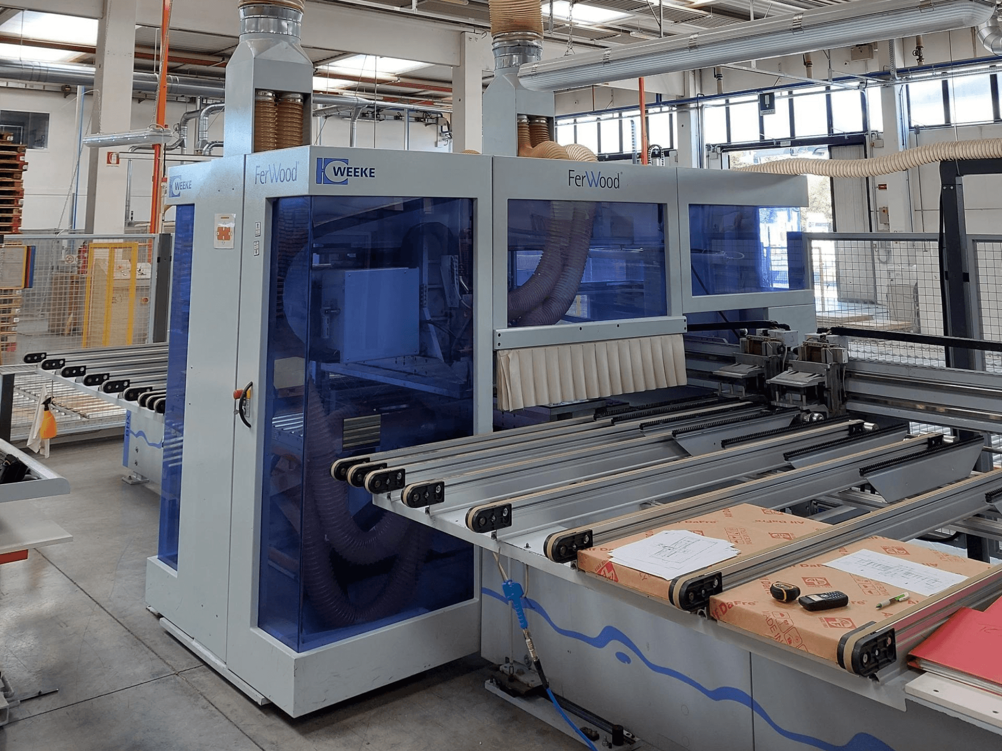 Weeke FerWood CNC makinesi bir atölyede, mavi renkli panelleri, kontrol düğmelerini ve malzemelerle çalışma masasını gösteren önden görünüm.