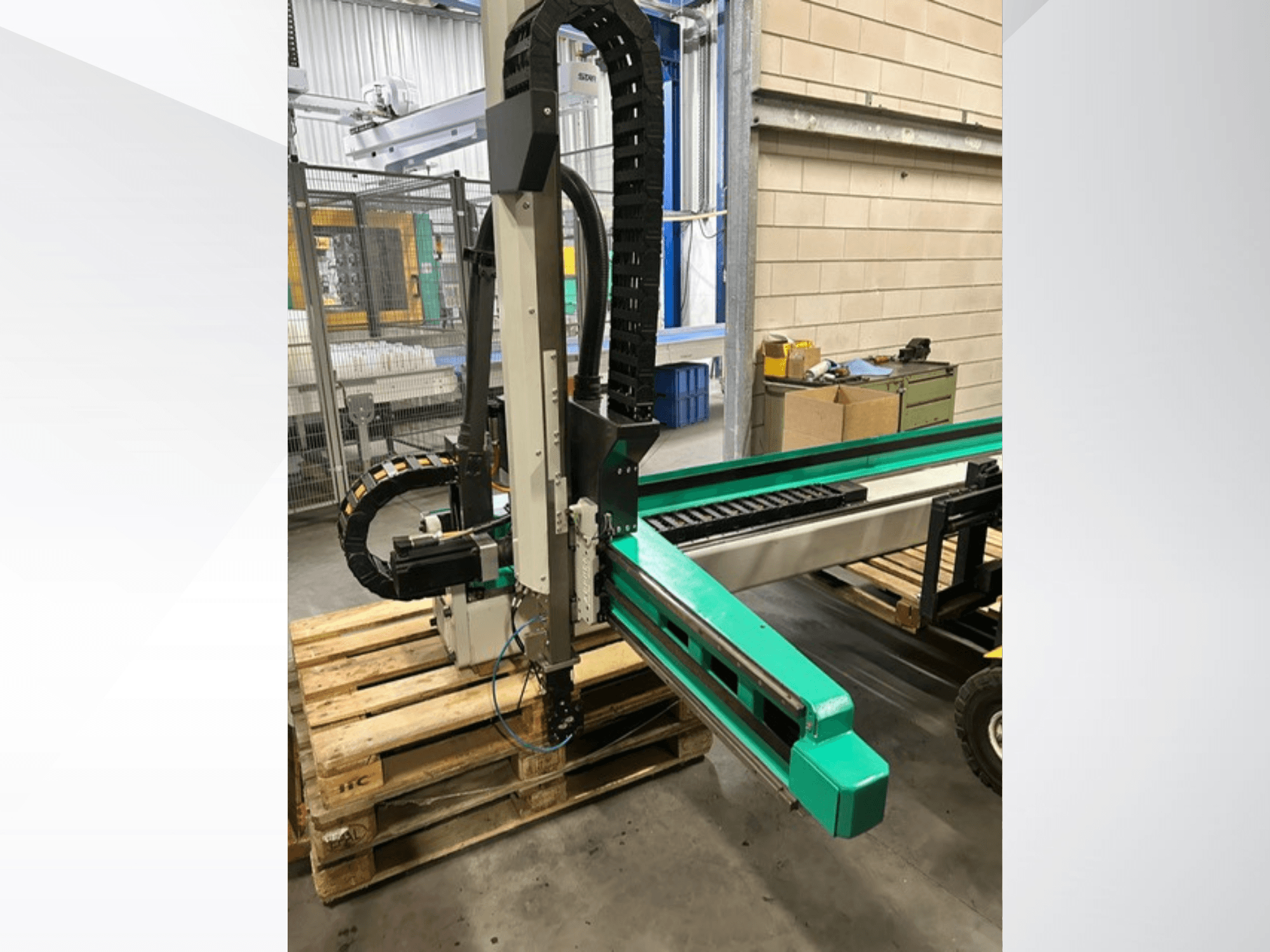 Arburg Multilift Select 15kg quer, ahşap paletler üzerinde iş parçası taşıma sisteminin yandan görünümü, robotik kol ve kontrol bileşenlerini gösterir.