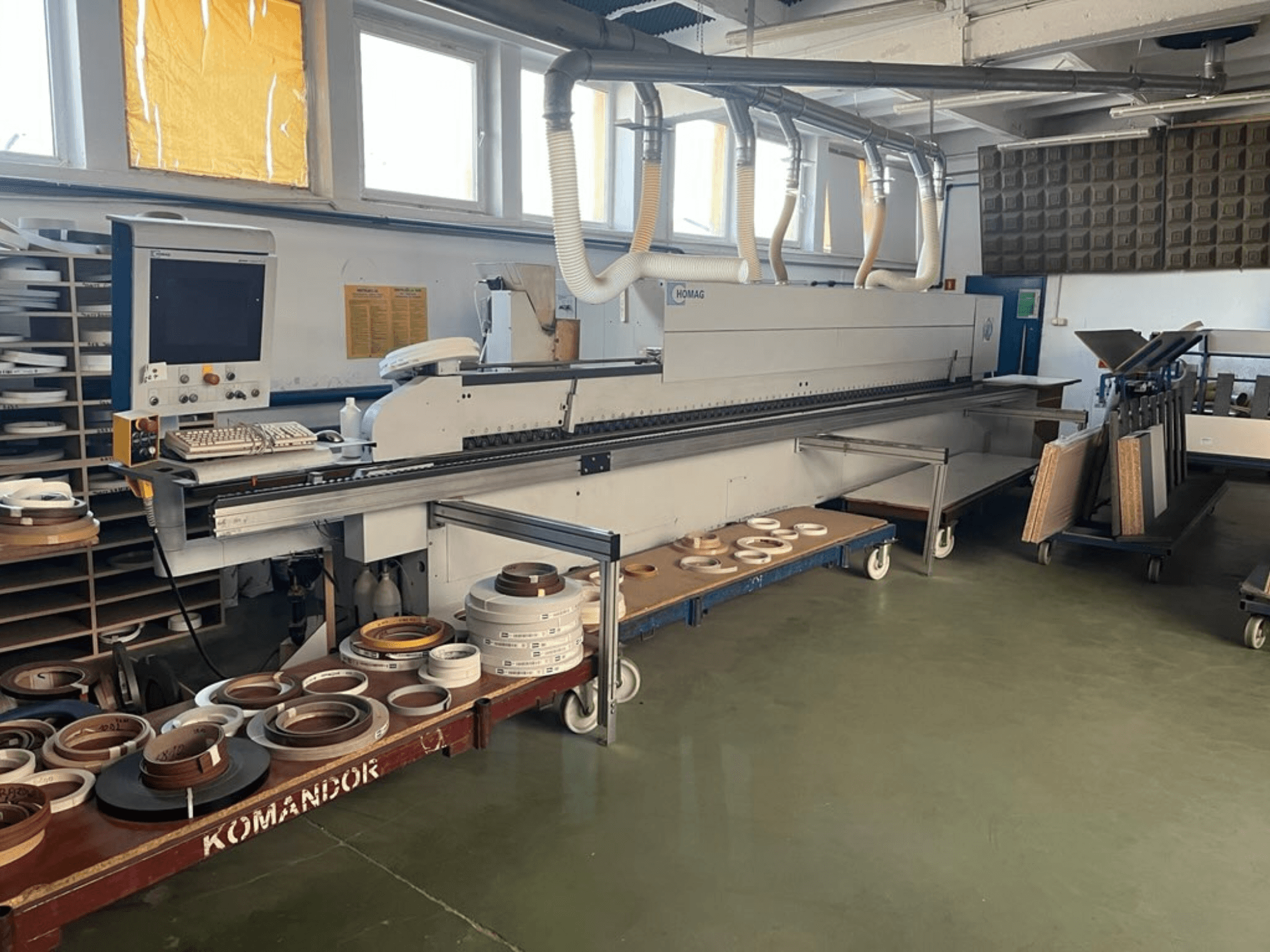 Atölyede HOMAG kenar bantlama makinesi, kumandalar ve mavi bir araba üzerinde çeşitli kenar bantlama malzemeleri. Yandan görünüm.