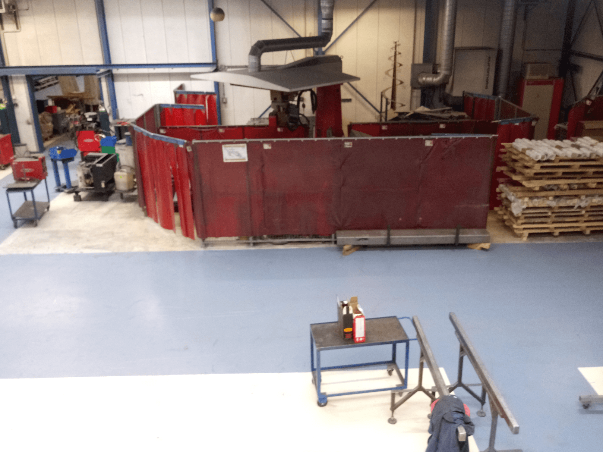 Kırmızı kaynak perdeleri, makineler ve paletler içeren endüstriyel çalışma alanı; düzen ve ekipmanı sergileyen üstten görünüm.