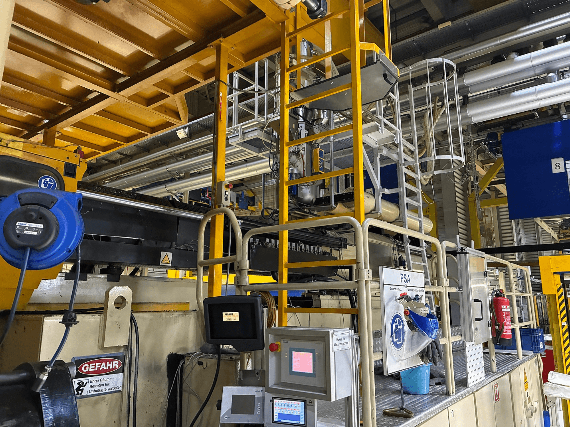 Mavi güvenlik ekipmanları ve sarı iskele ile endüstriyel makine kontrol paneli, boruları ve uyarı işaretlerini gösteren yandan görünüm.