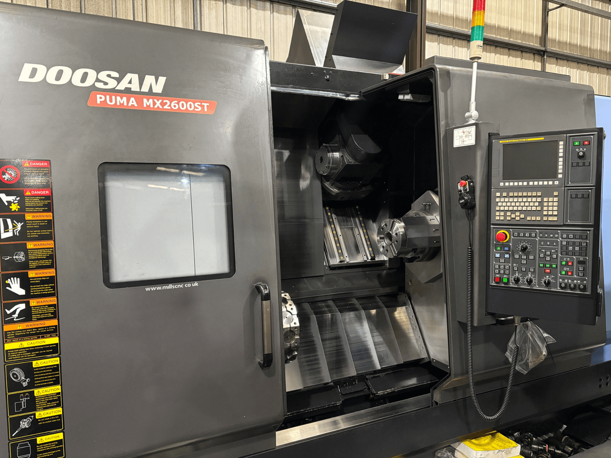 Doosan PUMA MX2600ST CNC torna tezgahı, önden görünüm, kontrol paneli, iş mili ve görünür güvenlik uyarıları ile işleme alanını gösterir.