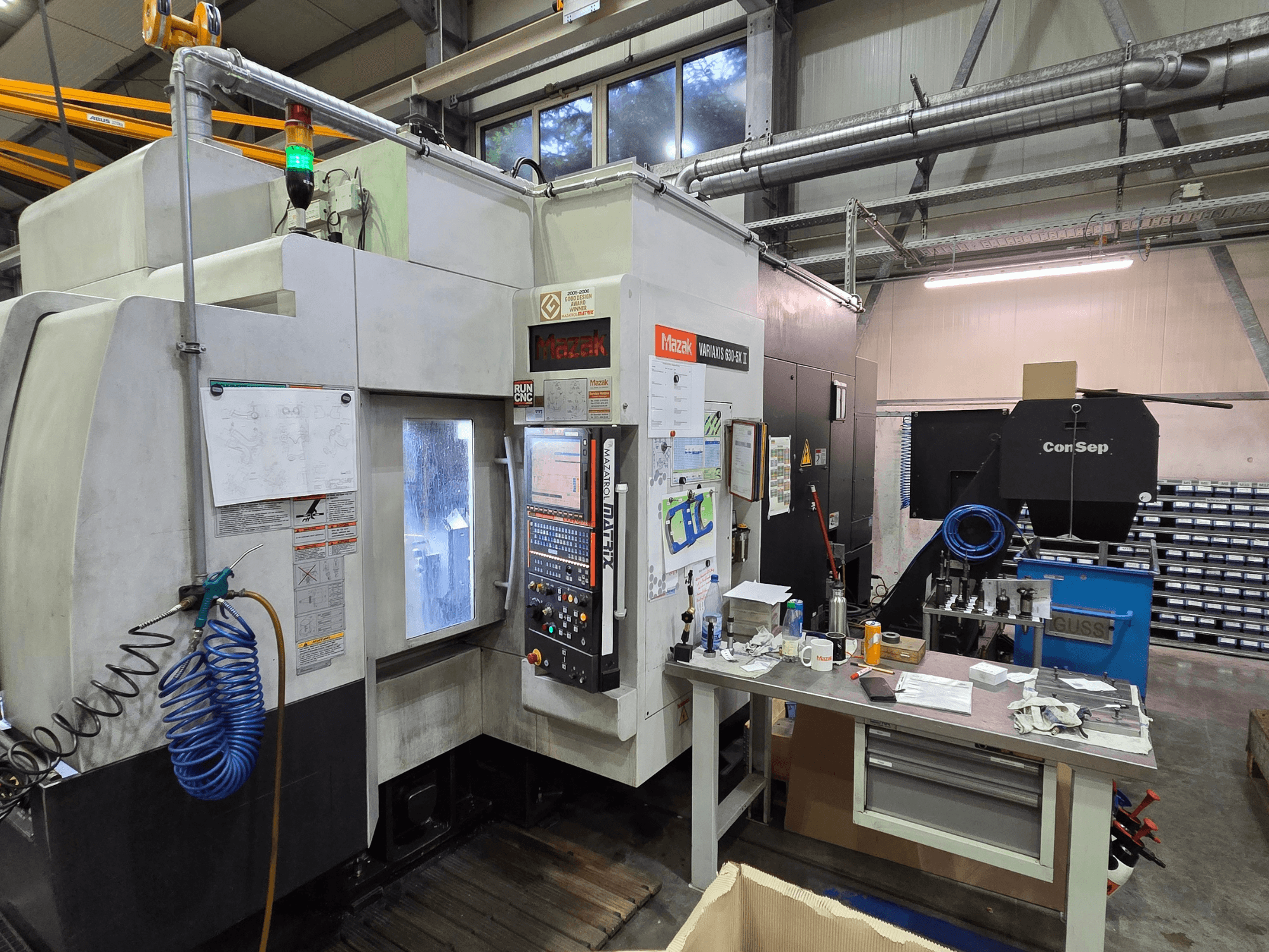 Atölyedeki Mazak Variaxis 630-5X CNC makinesi, kontrol paneli, hortum ve yan masadaki aksesuarlarla önden görünüm.