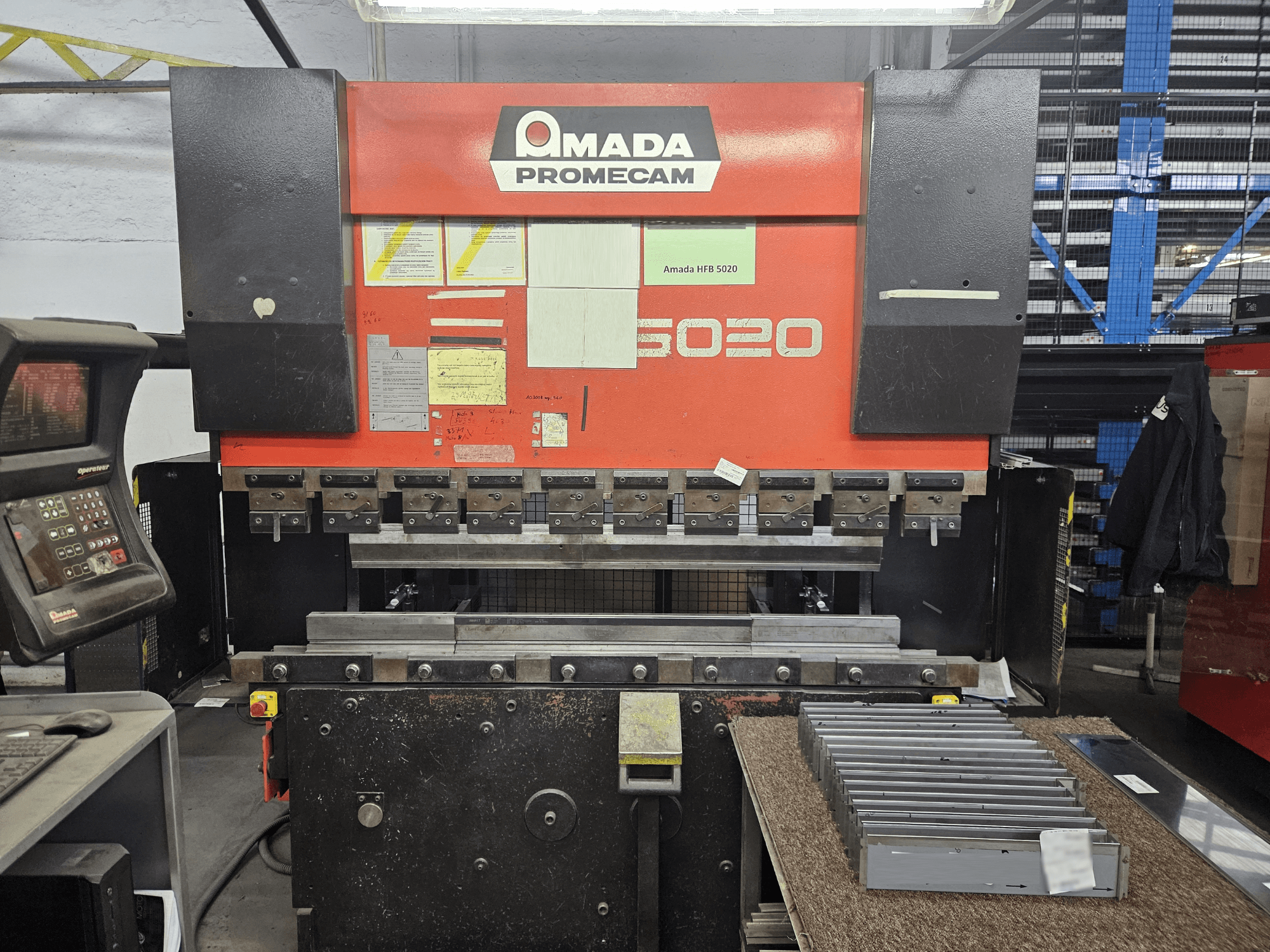 Amada HFBO 5020 abkant presin önden görünümü, kontrol paneli ve çalışma yüzeyinde istiflenmiş metal parçalar.