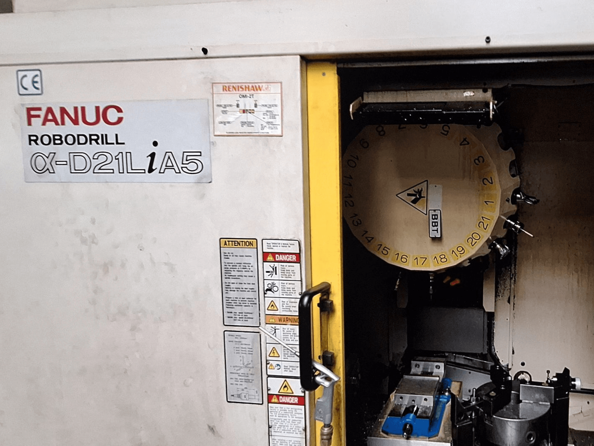 FANUC Robodrill α-D21LiA5, önden görünüm. Takım karuseli ve kelepçeli işleme alanını ve kapıdaki uyarıları gösterir.