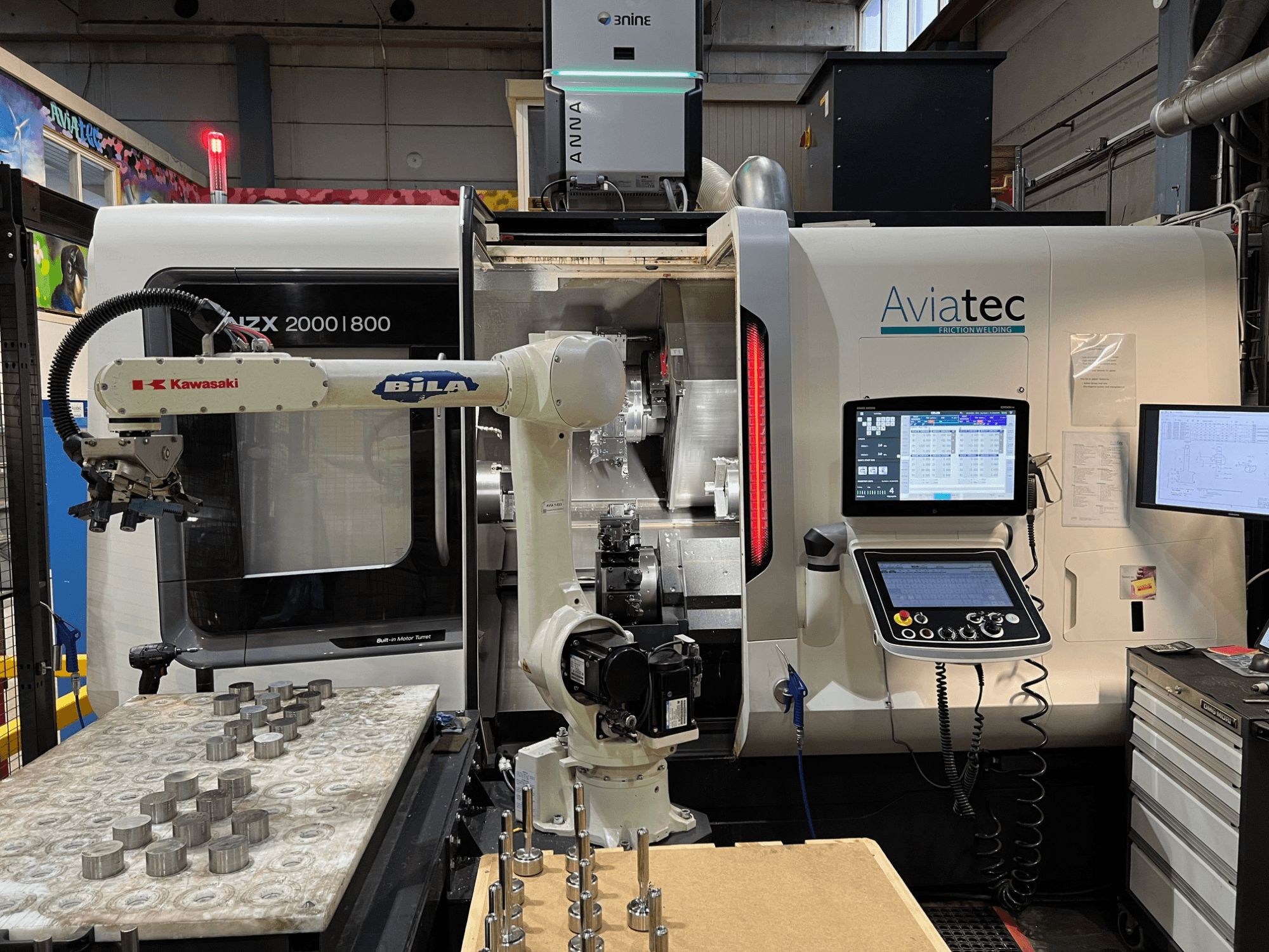 Kawasaki robot kolu, kontrol paneli ve çalışma masası üzerinde metal parçalar bulunan DMG MORI NZX 2000/800SY yatay torna tezgahı.