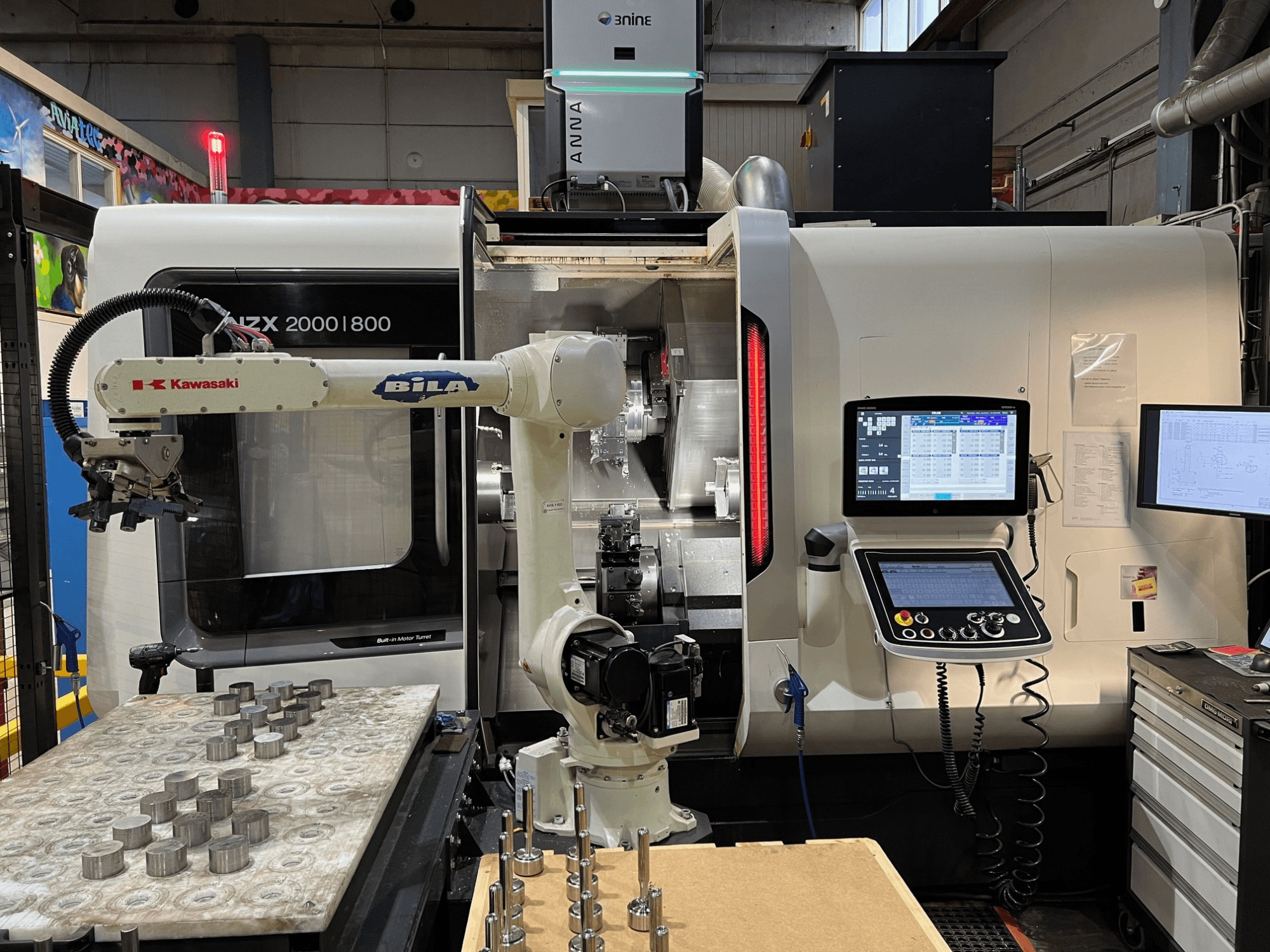 Bir atölyede bulunan DMG MORI NZX 2000/800SY yatay torna tezgahı; Kawasaki robot kolu ve kontrol panelleri görülebiliyor.