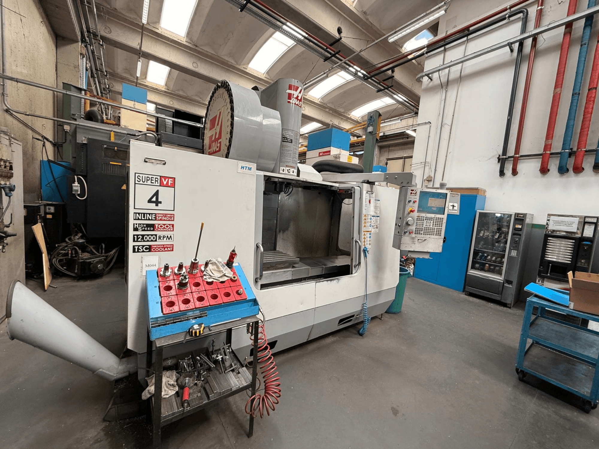 HAAS Super VF 4 CNC freze makinesi, kontrol paneli, 12.000 RPM iş mili ve mavi depolama rafı üzerinde takımlar, yandan görünüm.