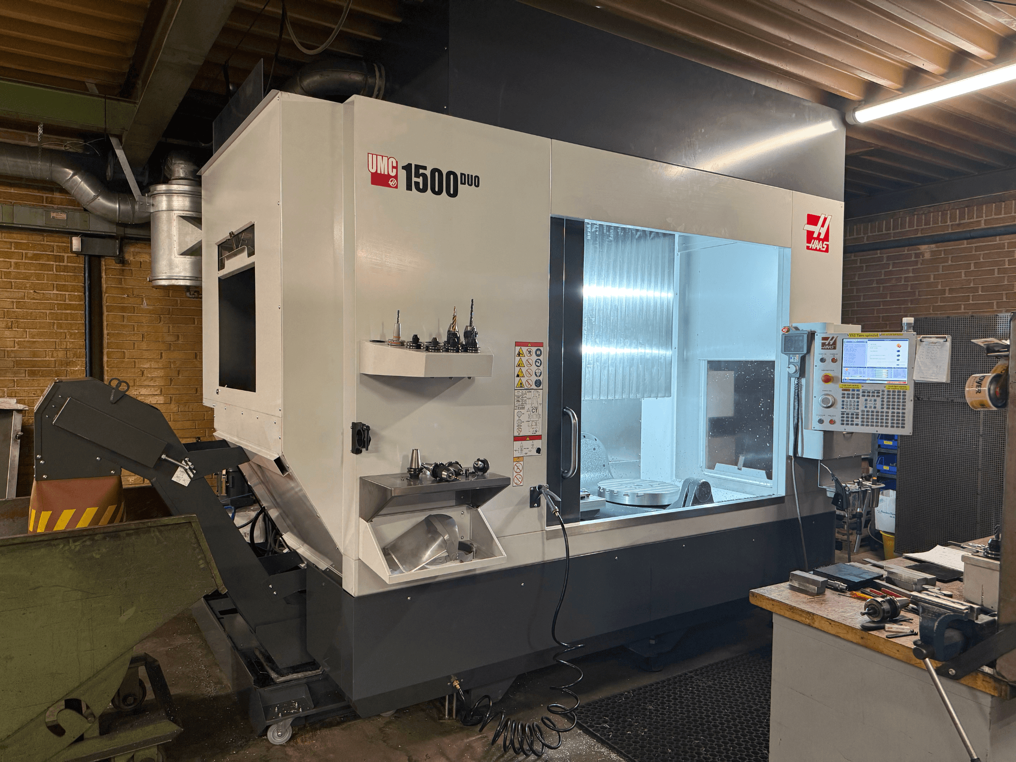 Haas UMC 1500DUO CNC makinesi, önden görünüm, büyük bir pencere, kontrol paneli ve bir raf üzerinde takım tutuculara sahiptir.