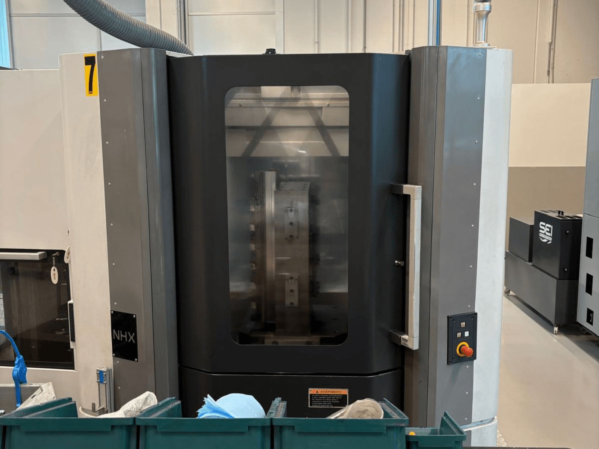 NHX CNC makinesi önden görünümde, kontrol panelini ve net ekranı gösterir; yakınında yeşil kutular bulunan bir atölyede bulunur.