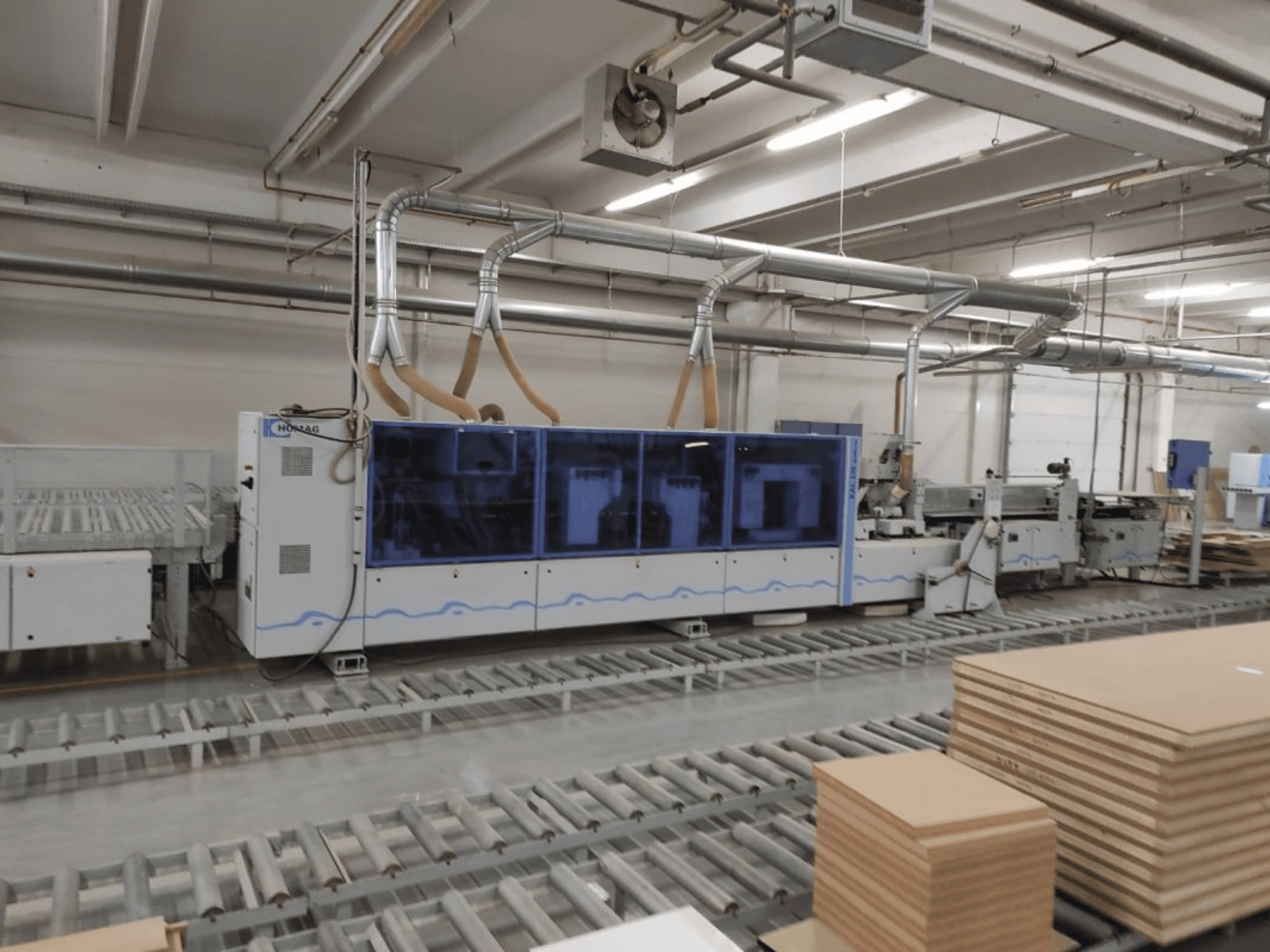 Bir fabrikadaki HOMAG üretim makinesi, önden görünüm, mavi kasa ve istiflenmiş kontrplak levhalar içeren konveyör sistemi.