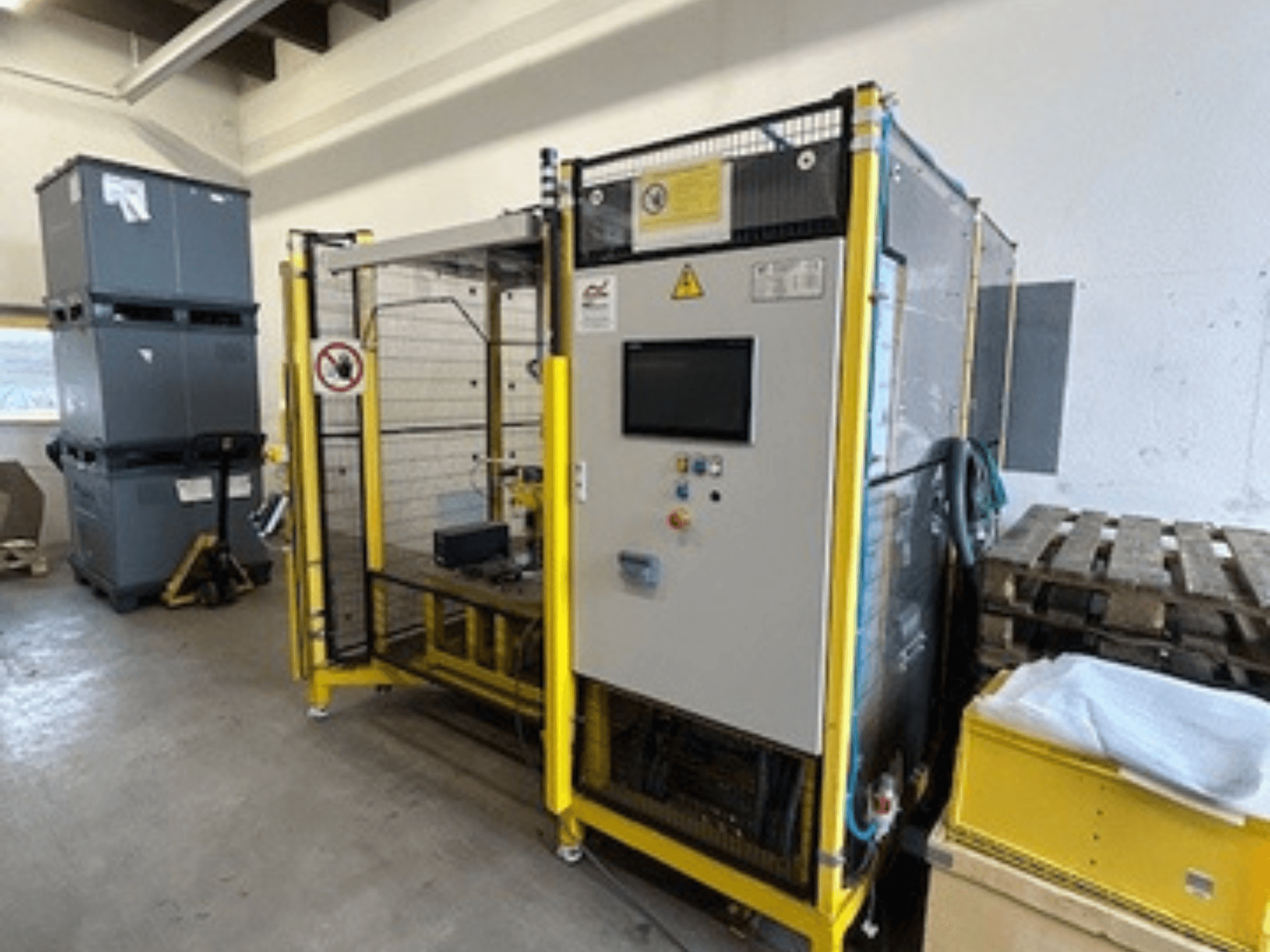 Kontrol paneli ve güvenlik özellikleri olan endüstriyel makine, önden görünüm; arka planda sarı çerçeve, gri kutular ve paletler.