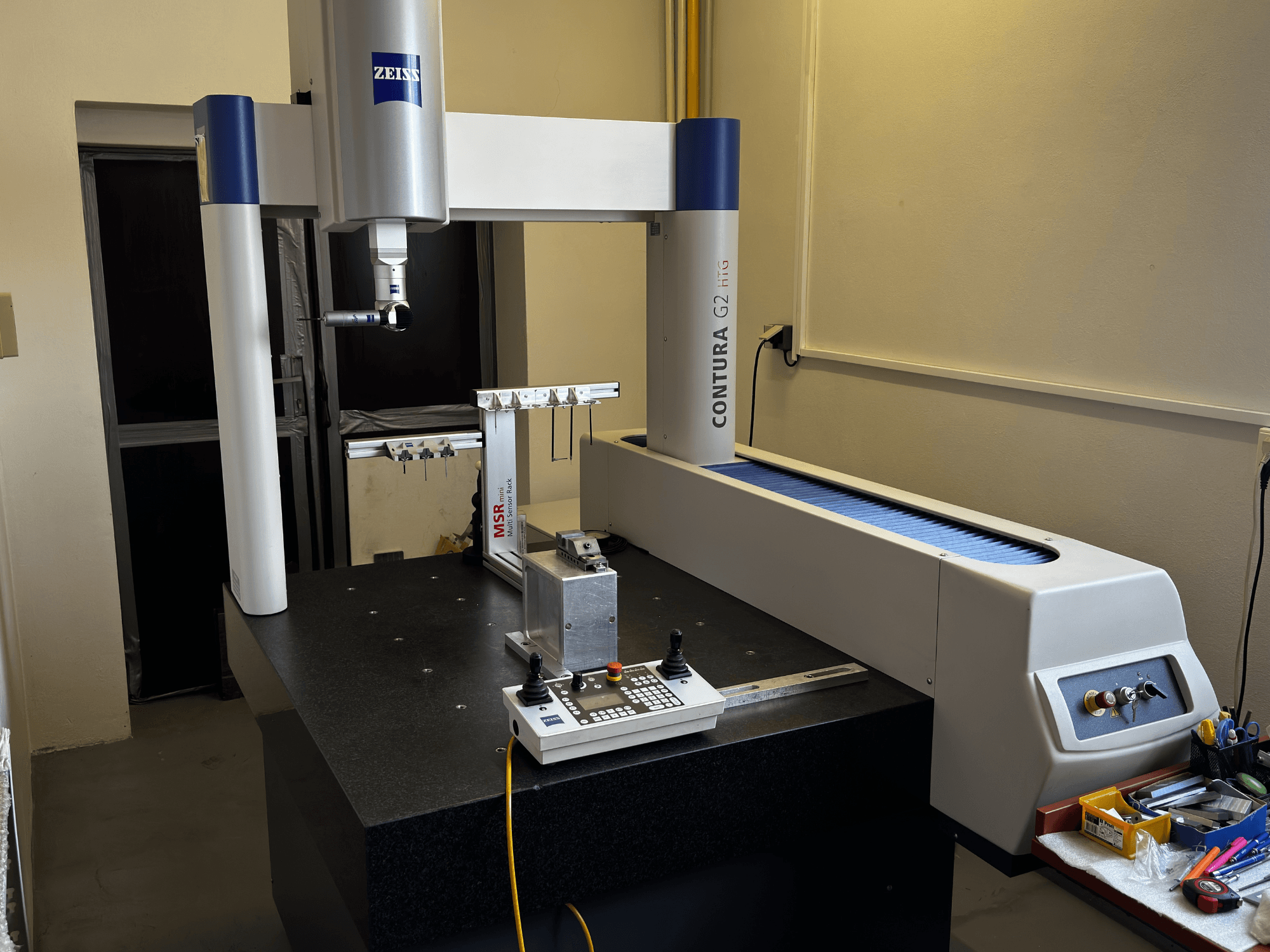 Laboratuvar ortamında ZEISS Contura G2 CMM, ölçüm kolu, kontrol paneli ve bitişik çalışma alanını gösteren önden görünüm.