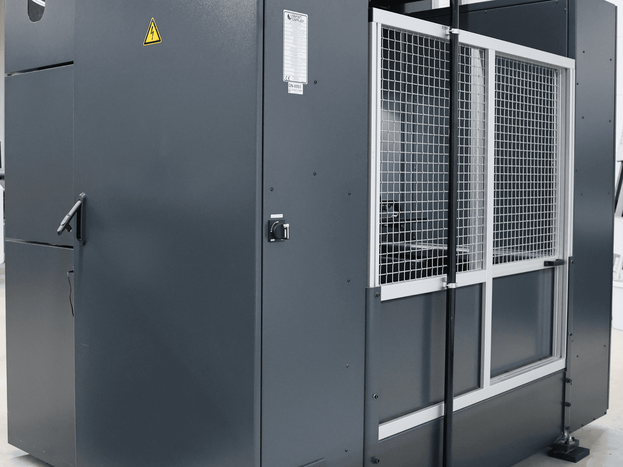 Güvenlik uyarılı gri endüstriyel makine, kafesli kapı ve kontrol panelini gösteren önden görünüm, model etiketli.