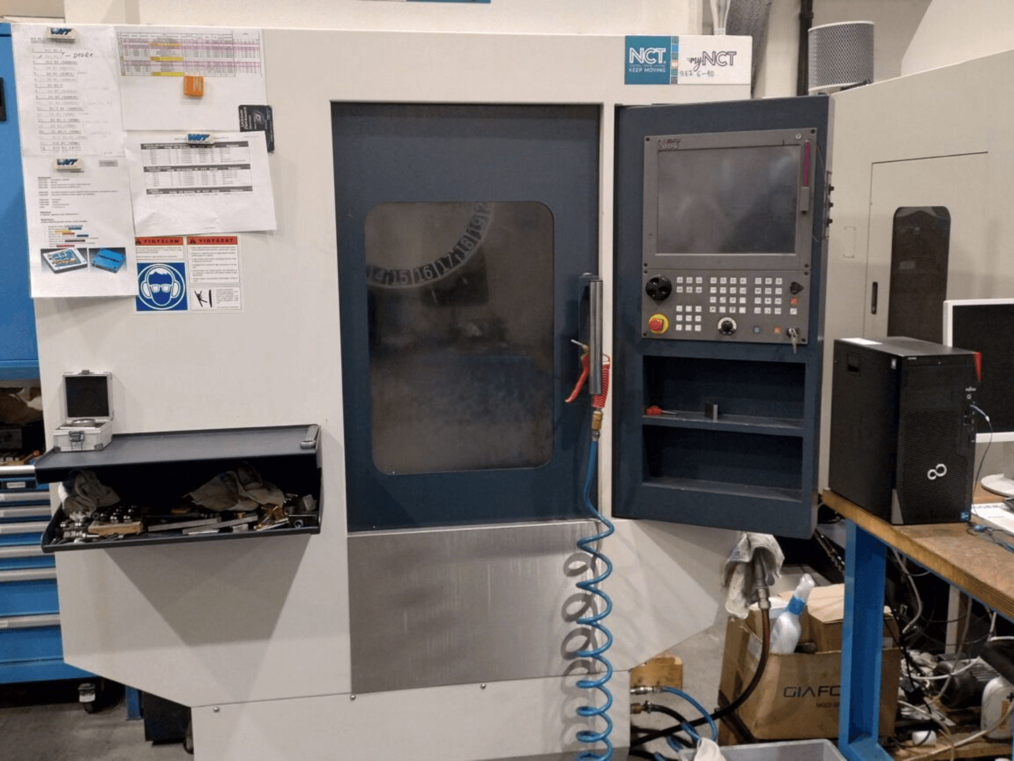 NCT CNC makinesi, önden görünüm, sayısal tuş takımlı kontrol paneli, saklama çekmecesinde aletler ve duvarda kılavuz bulunmaktadır.