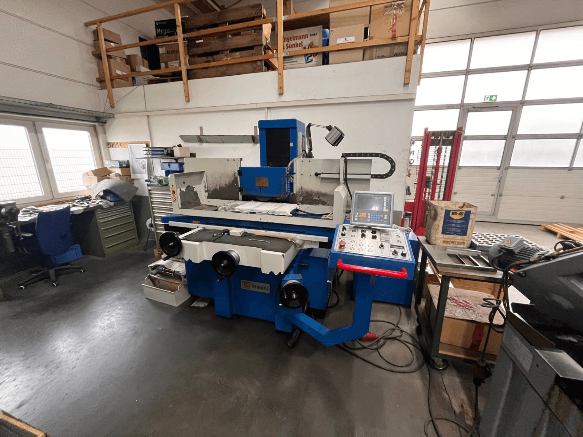 Dijital kontrol panelli mavi yüzey taşlama makinesi, önden görünüm, çalışma alanındaki dağınıklığı ve yeşil bir alet dolabını gösteriyor.