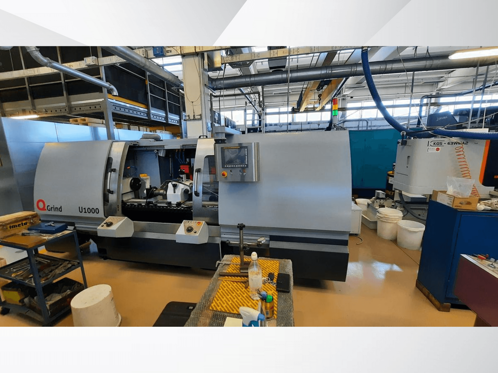 Q-Grind U1000 makinesi bir atölyede, kontrol paneli ve açık kesim alanı ile önden görünüm, ekipmanla çevrili.