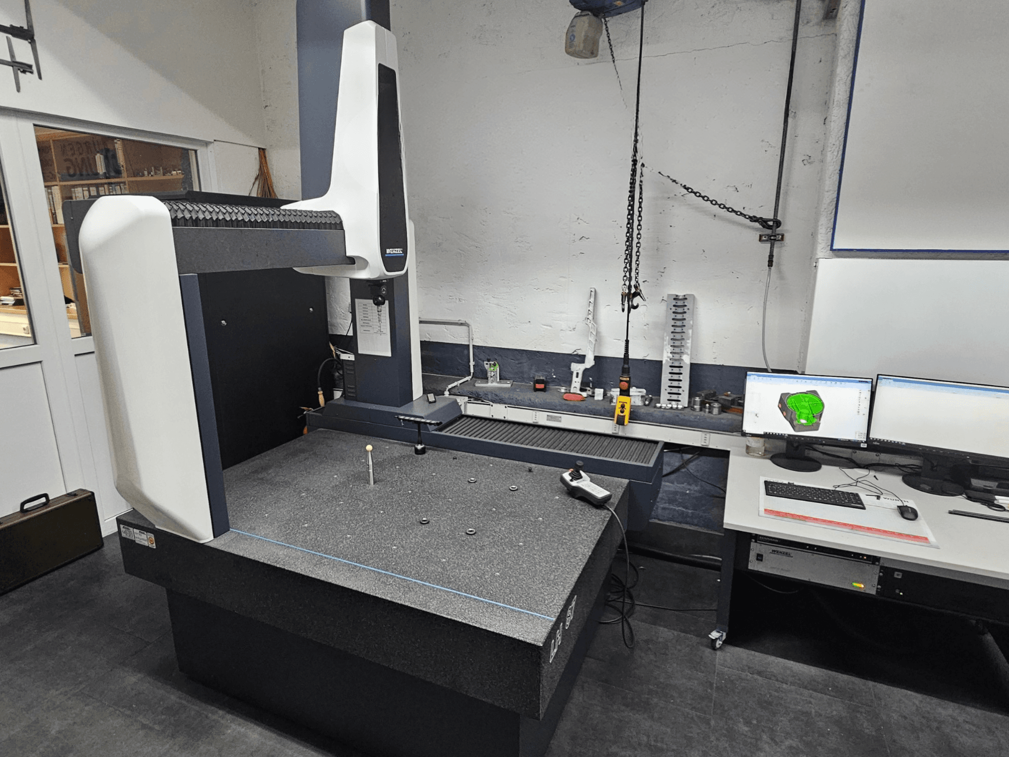 Koordinat Ölçüm Makinesi Wenzel LH 87 bir atölyede, önden görünüm, granit tabla üzerinde bir prob ve CAD tasarımını gösteren çift monitör.