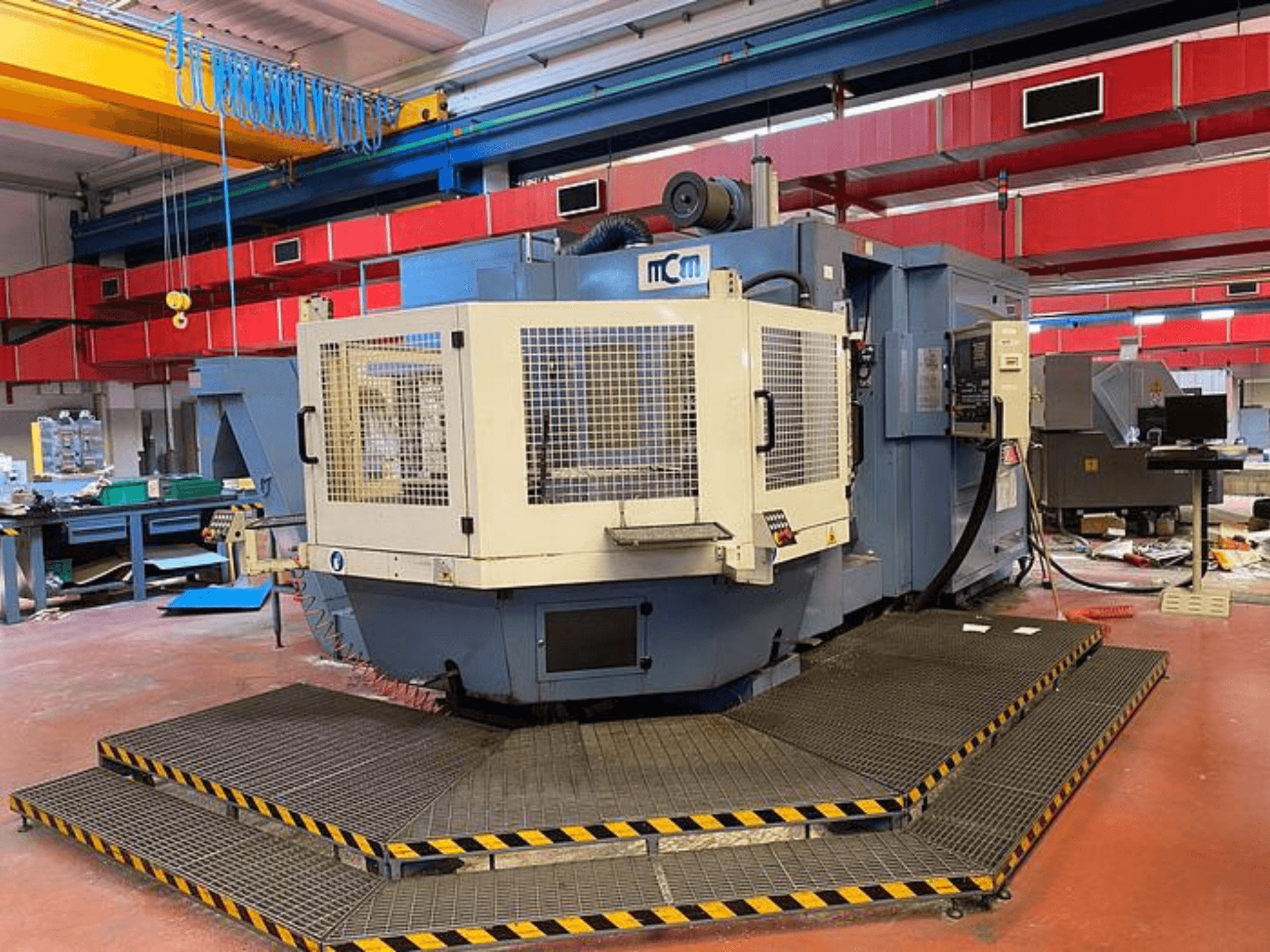 Bir fabrikadaki MCM CNC makinesi, koruyucu kafesli önden görünüm, siyah ve sarı güvenlik kenarları, endüstriyel ekipmanlarla çevrili.