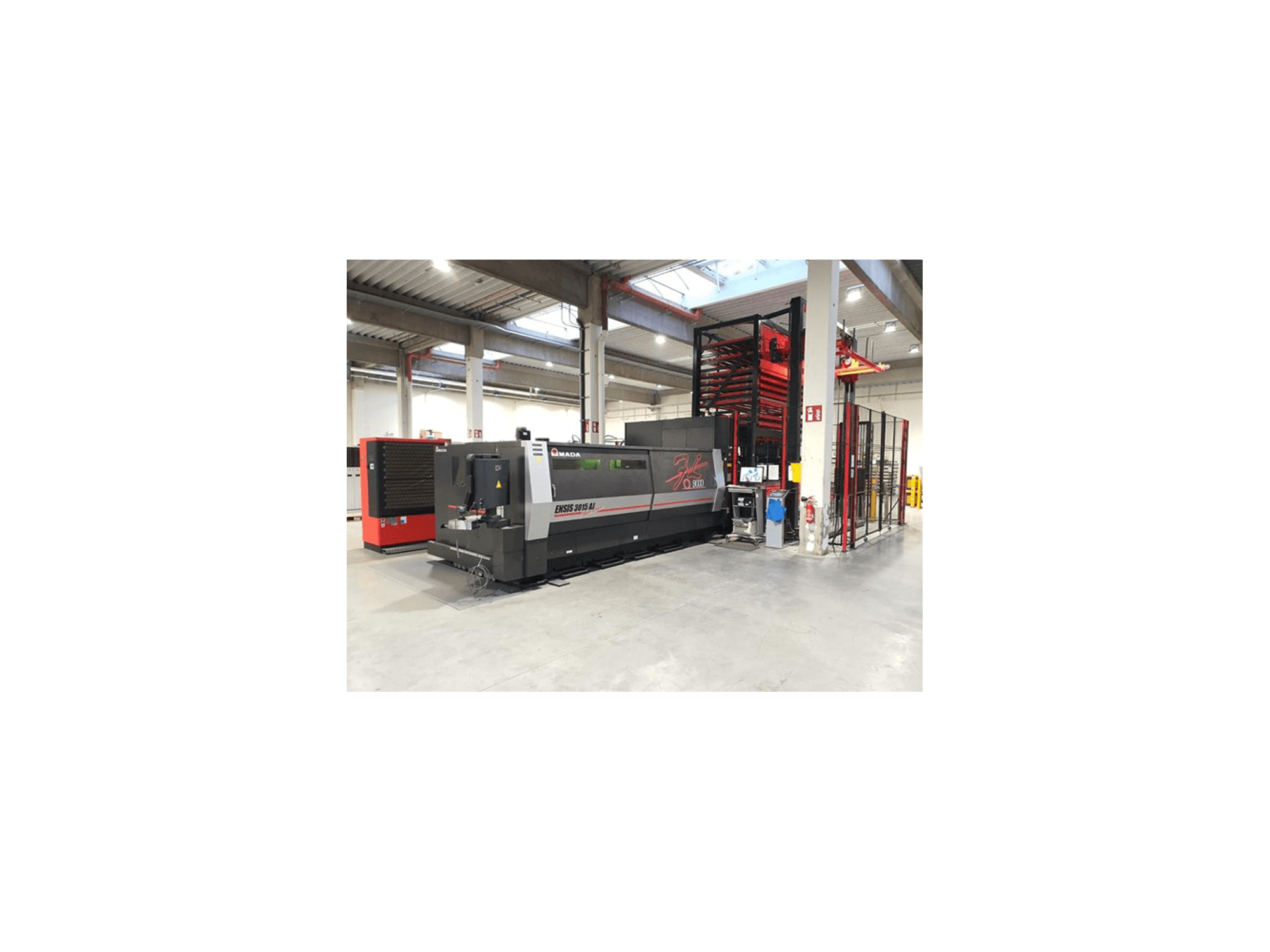 Amada ENSIS 3015 Aj lazer kesim makinesinin önden görünümü, metal raflı geniş bir endüstriyel atölyeye yerleştirilmiştir.