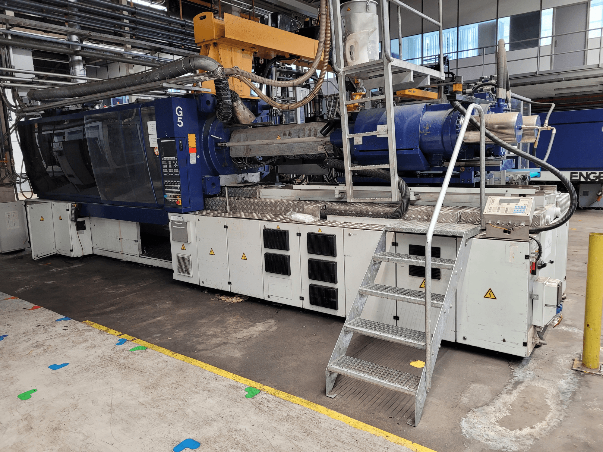 Enjeksiyon kalıplama makinesi G5, yandan görünüm, kontrol paneli, basamaklı merdiven ve paslanmaz çelik çalışma yüzeyi görünür.