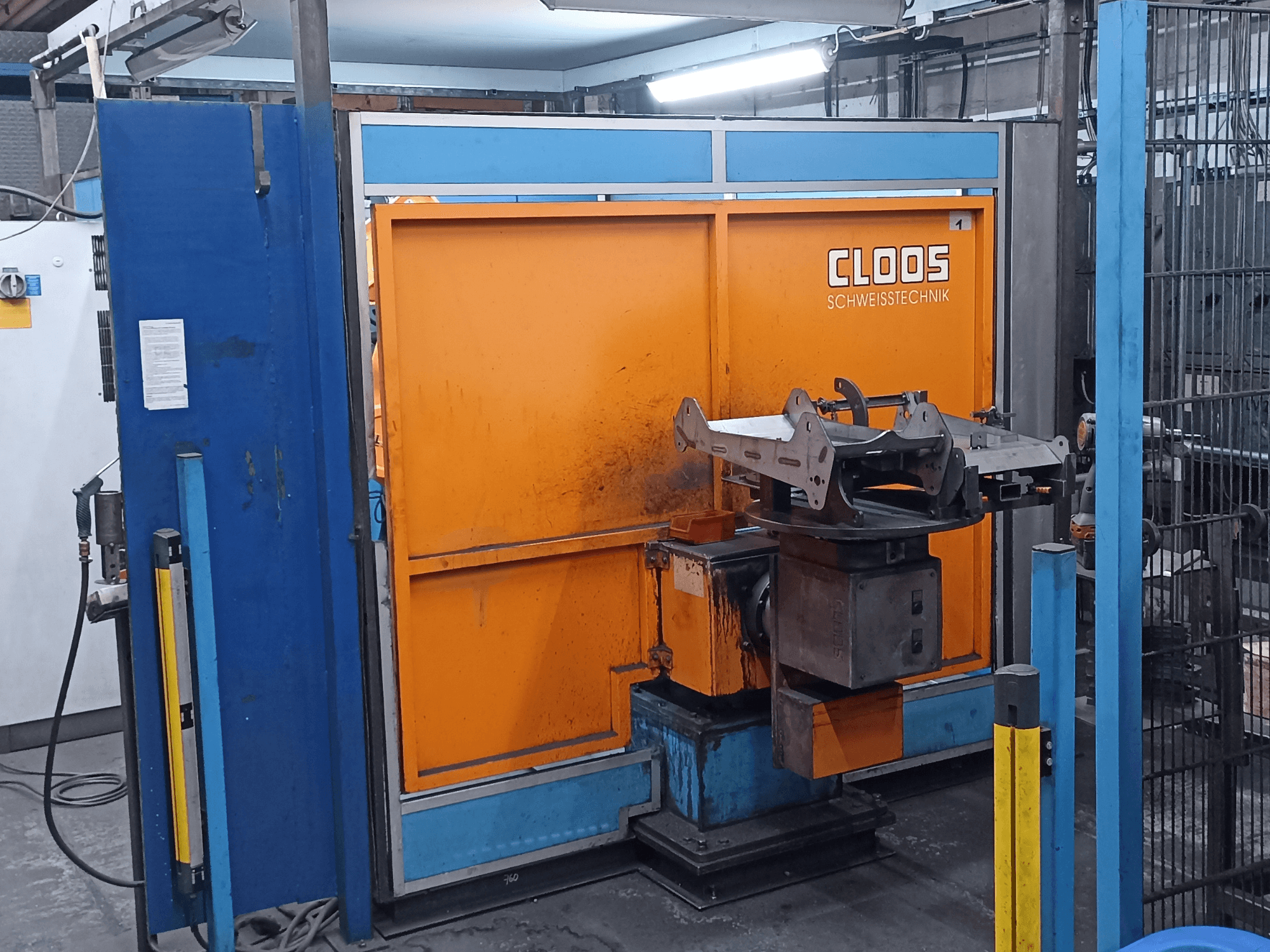 Bir atölyede bulunan CLOOS Romat 320 kaynak robotu, önden görünüm; turuncu bir kontrol paneli ve döner bir taban üzerinde metal bir iş parçası ile.
