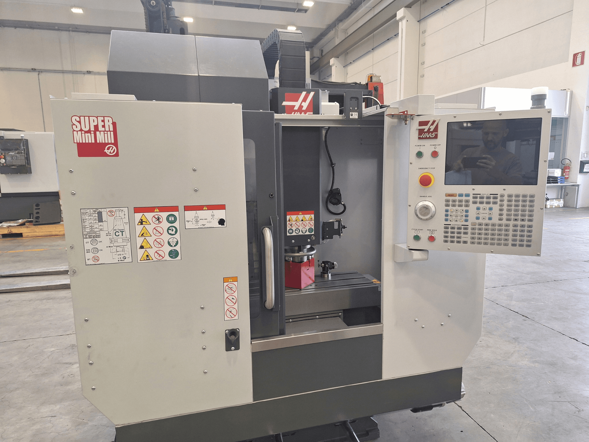 HAAS Super Mini Mill’in ön görünümü; kontrol paneli, güvenlik etiketleri ve iş parçası yerleştirilmiş işleme alanını göstermektedir.