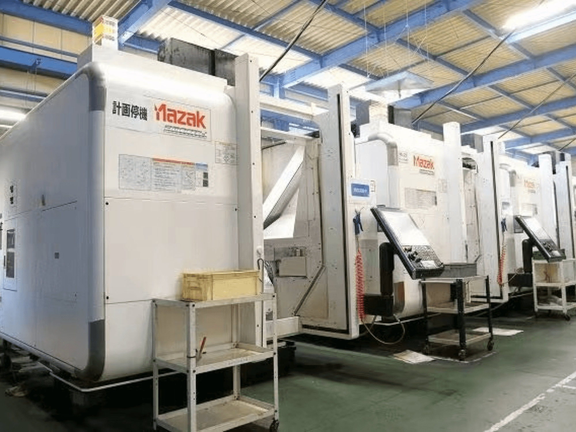 Fabrika ortamında Mazak CNC makineleri, kontrol panellerini ve beyaz gövdeler üzerindeki etiketleri gösteren yandan görünüm.