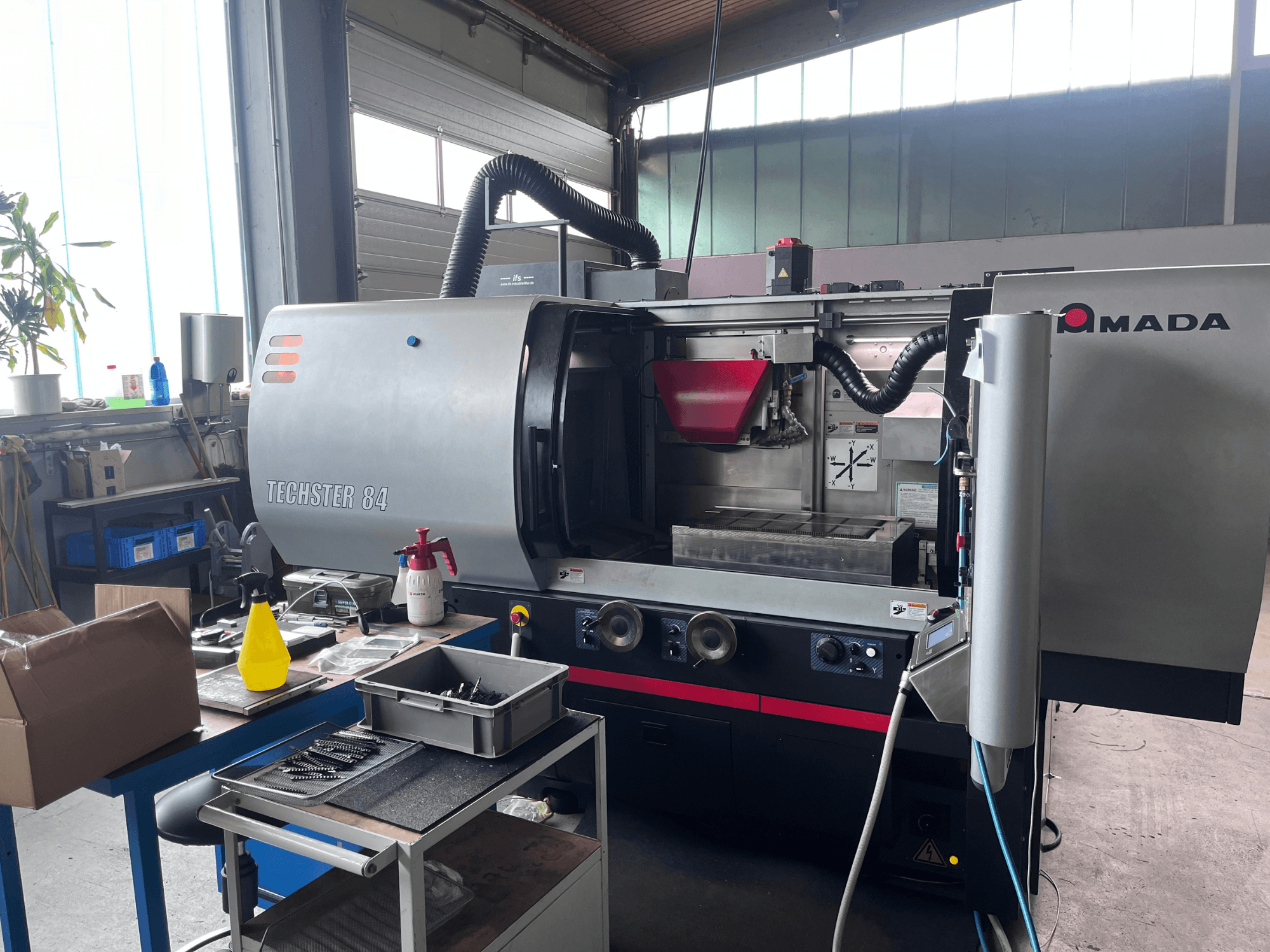 Bir atölyede bulunan AMADA Techster 84 yüzey taşlama makinesi; ön görünümünde çalıştırma kontrolleri ve kırmızı besleme haznesi görülüyor.