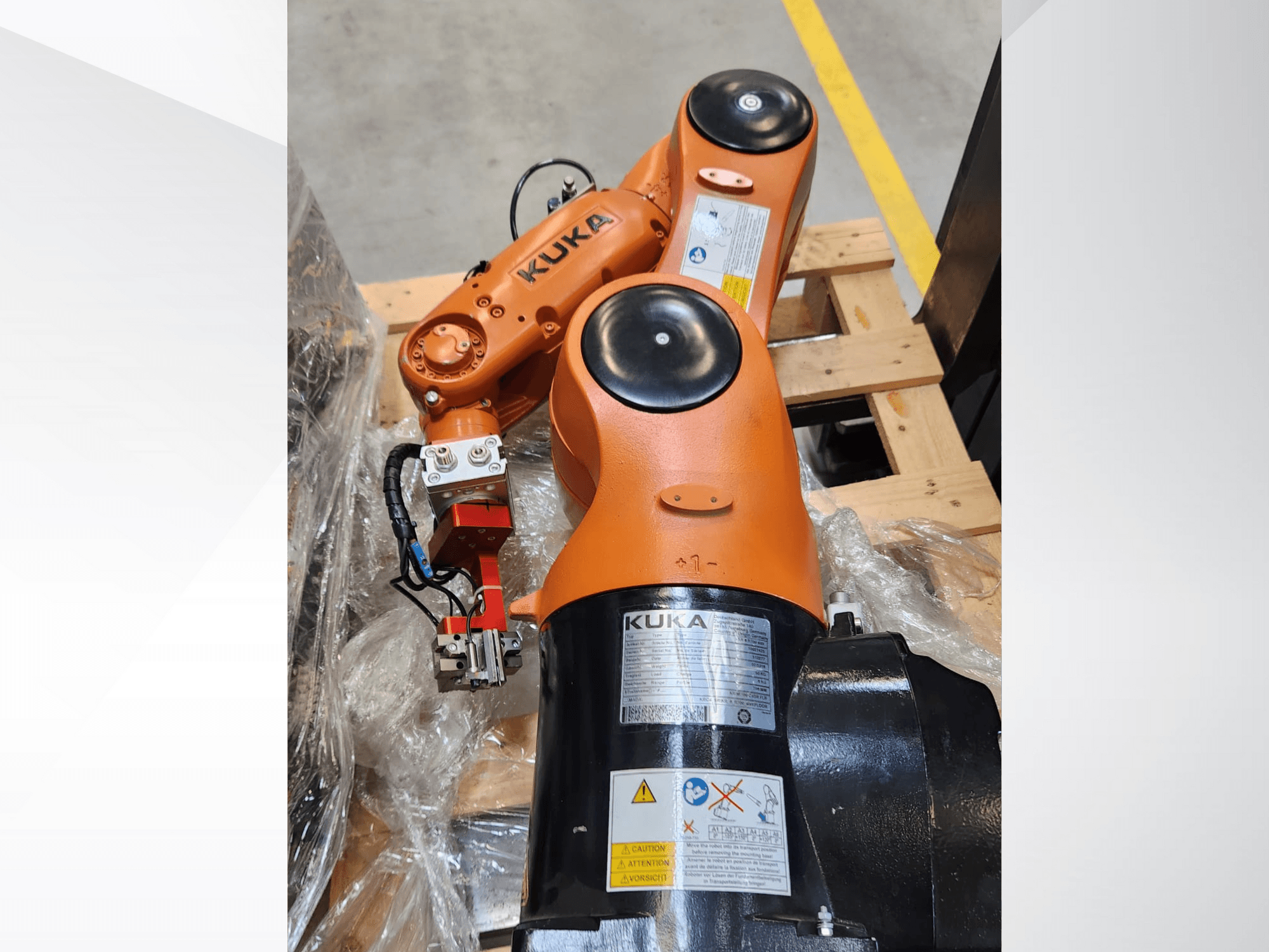 Turuncu kaplamalı KUKA robot kolu, yandan görünüm, ahşap bir palet üzerinde bir kavrayıcı ve kontrol kablolarına sahip.