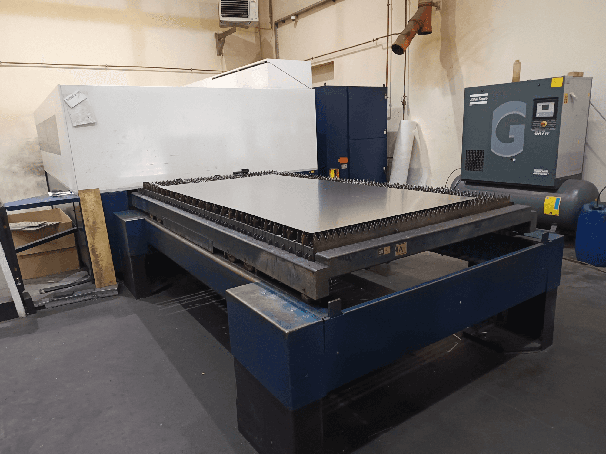 Metal tablalı endüstriyel kesim makinesi, önden görünüm. Keskin kenarlar, sağda hava kompresörü ve çelik çerçeve.
