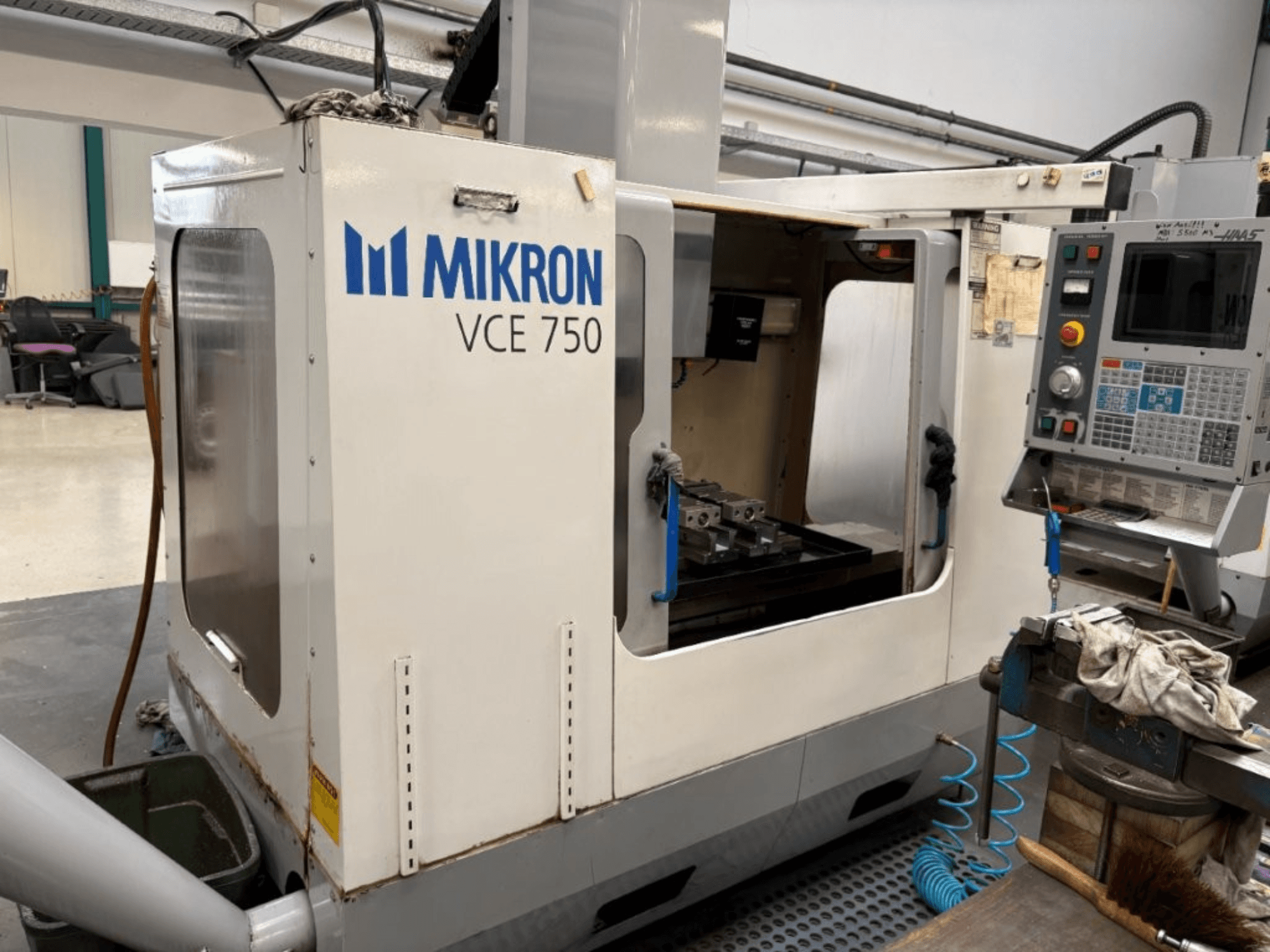 Mikron VCE 750 CNC freze makinesinin önden görünümü, işlenmiş bir çalışma tezgahı ve çeşitli düğmeler ve ekranlar içeren kontrol paneli.