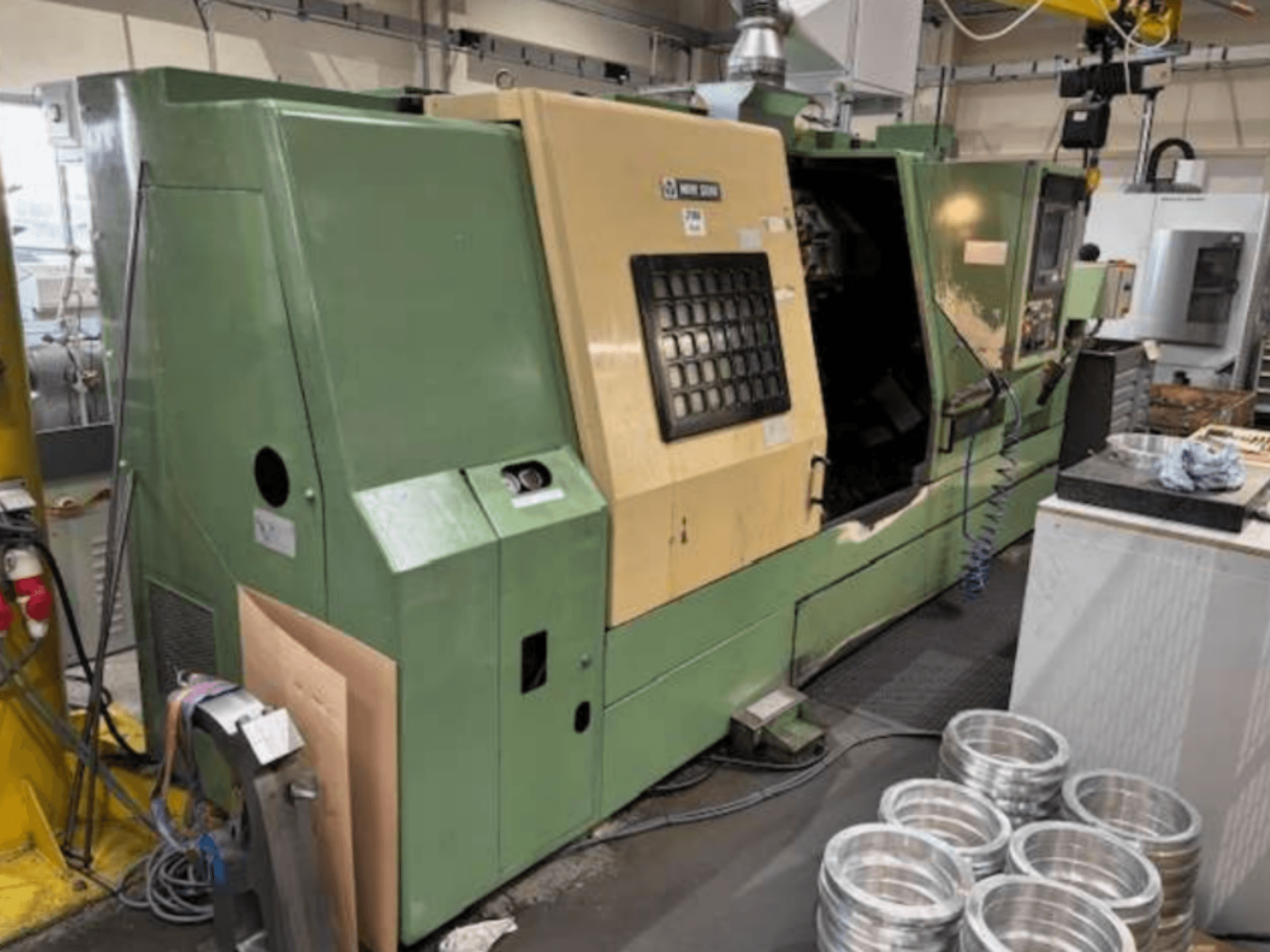 Mazak'ın yeşil CNC torna makinesi, önden bakıldığında bir kontrol paneli ve çalışma tezgahı üzerinde işlenmiş alüminyum parçalar sergiliyor.