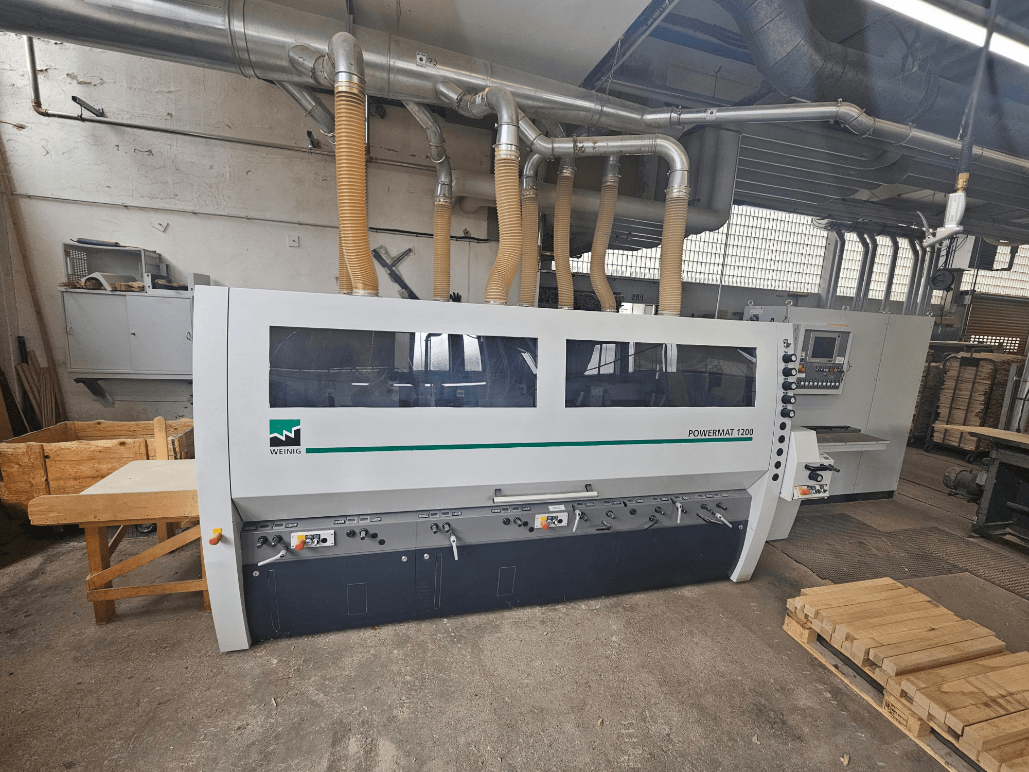 Weinig Powermat 1200 ahşap işleme makinesi, önden görünüm, kontrol paneli ve atölye ortamında ahşap yığınları ile.