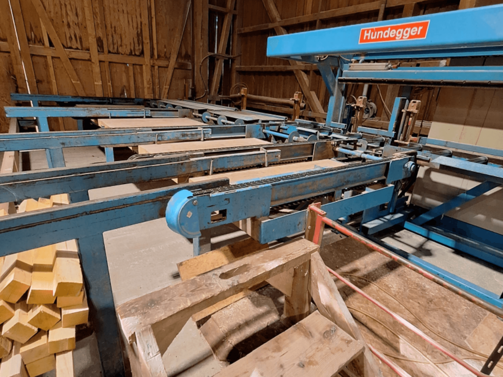 Bir ahırda bulunan, mavi metal iskelet, konveyör sistemi ve üst üste dizilmiş ahşap kirişlere sahip HUNDEGGER K2-A CNC Ahşap İşleme Merkezi.