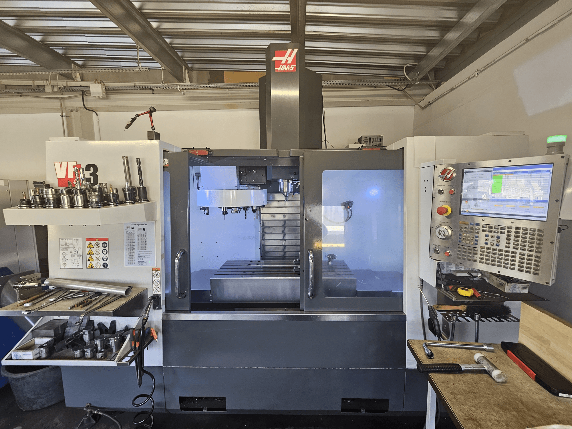 Atölyede Haas VF-3 CNC makinesi, önden görünümde aletler, kontroller ve aydınlatılmış iç kısım ile frezeleme alanı.