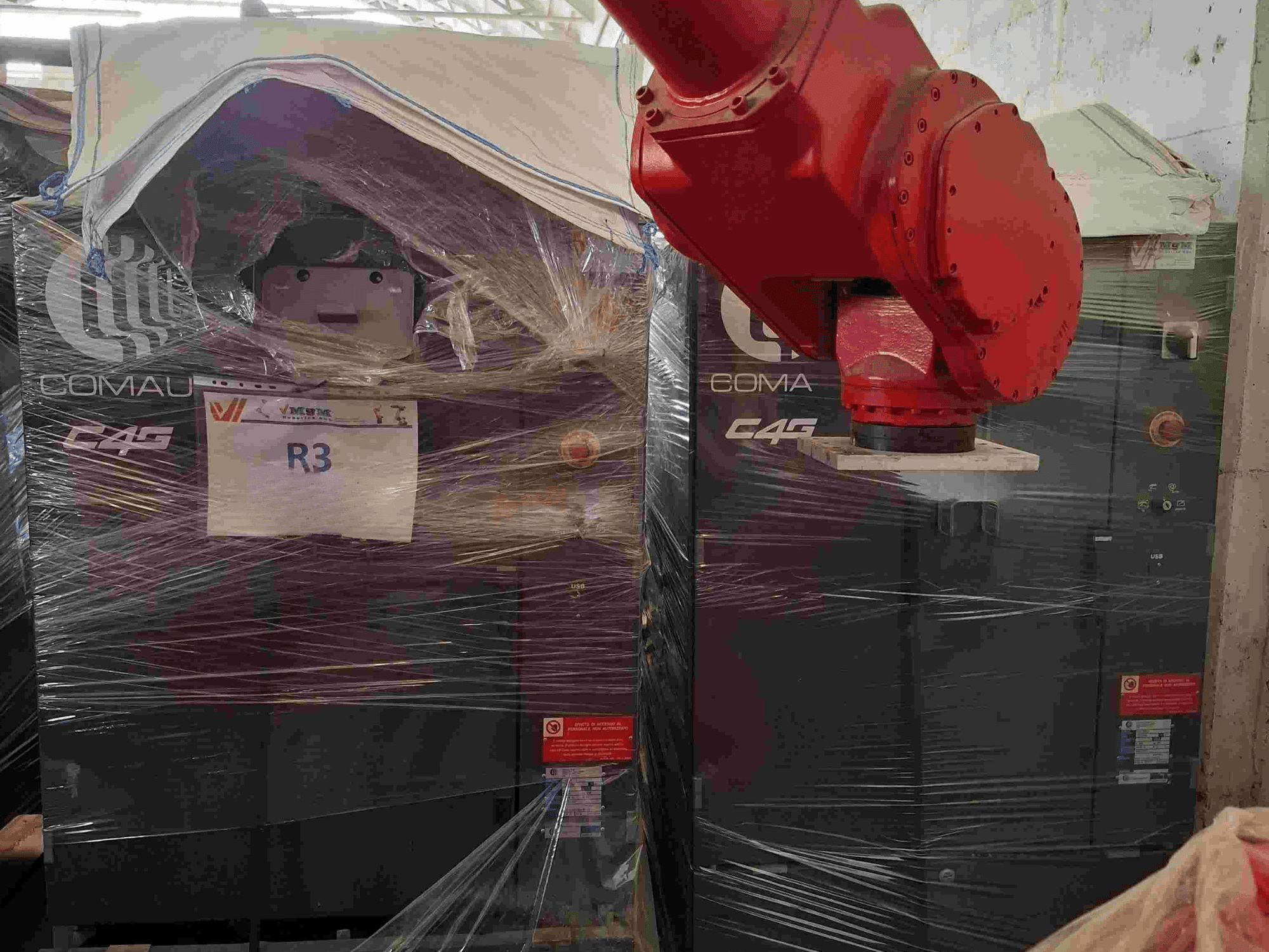 Comau C4G robot kolu önden görülüyor, plastiğe sarılmış, "R3" etiketli, makinenin üzerinde uyarı etiketleri görülüyor.